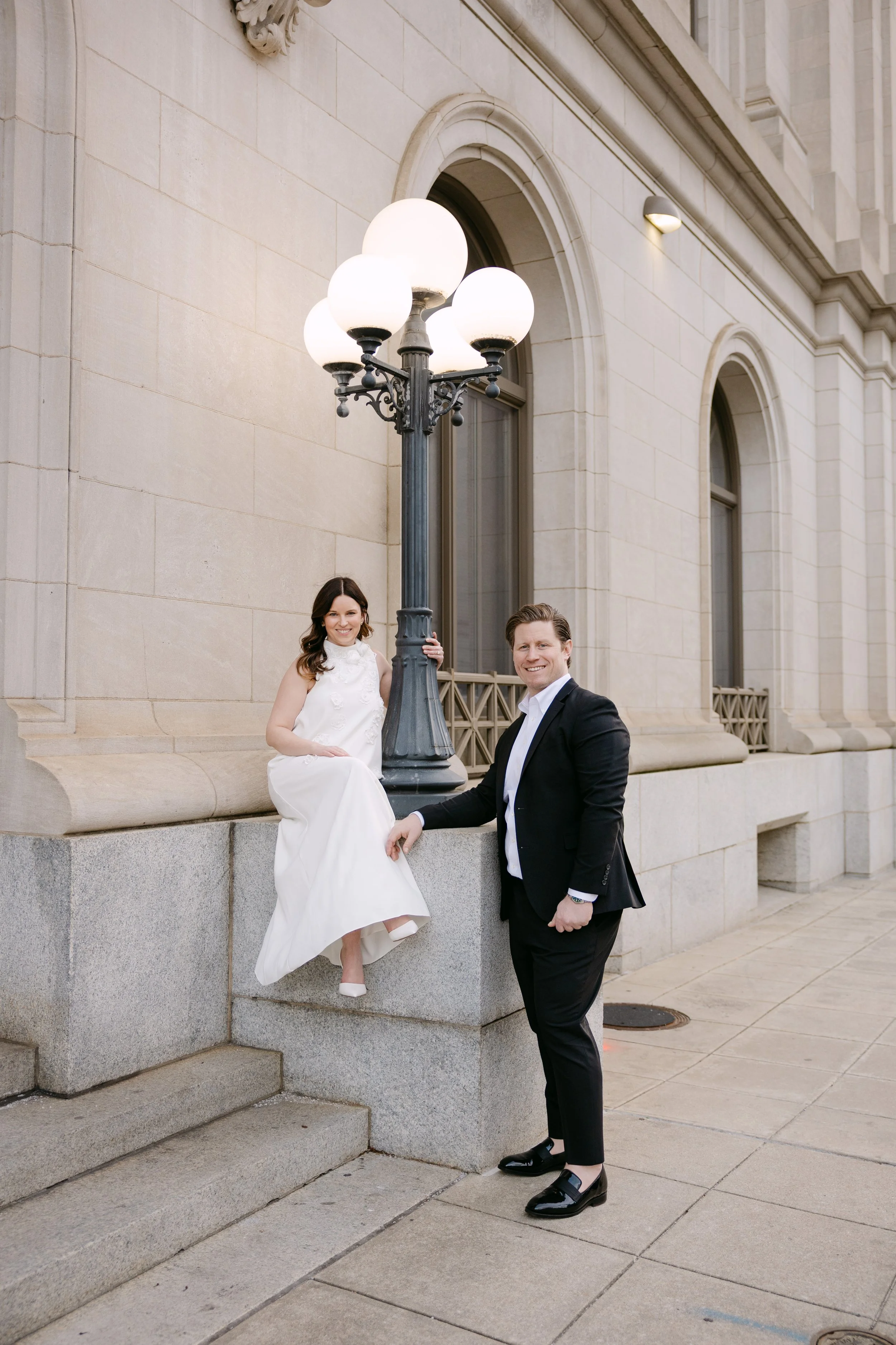 A woman in a white dress sitting on a stone ledge next to a man in a black suit standing outdoors in front of an elegant historic building with large arched windows and a decorative streetlamp.