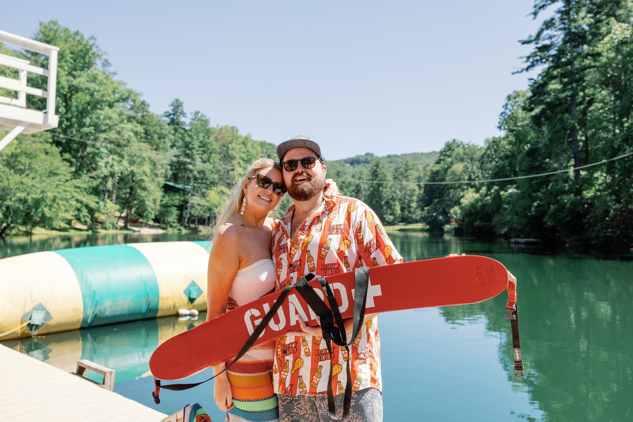 A smiling couple standing on a dock by a lake, holding a red rescue float, surrounded by trees on a sunny day.