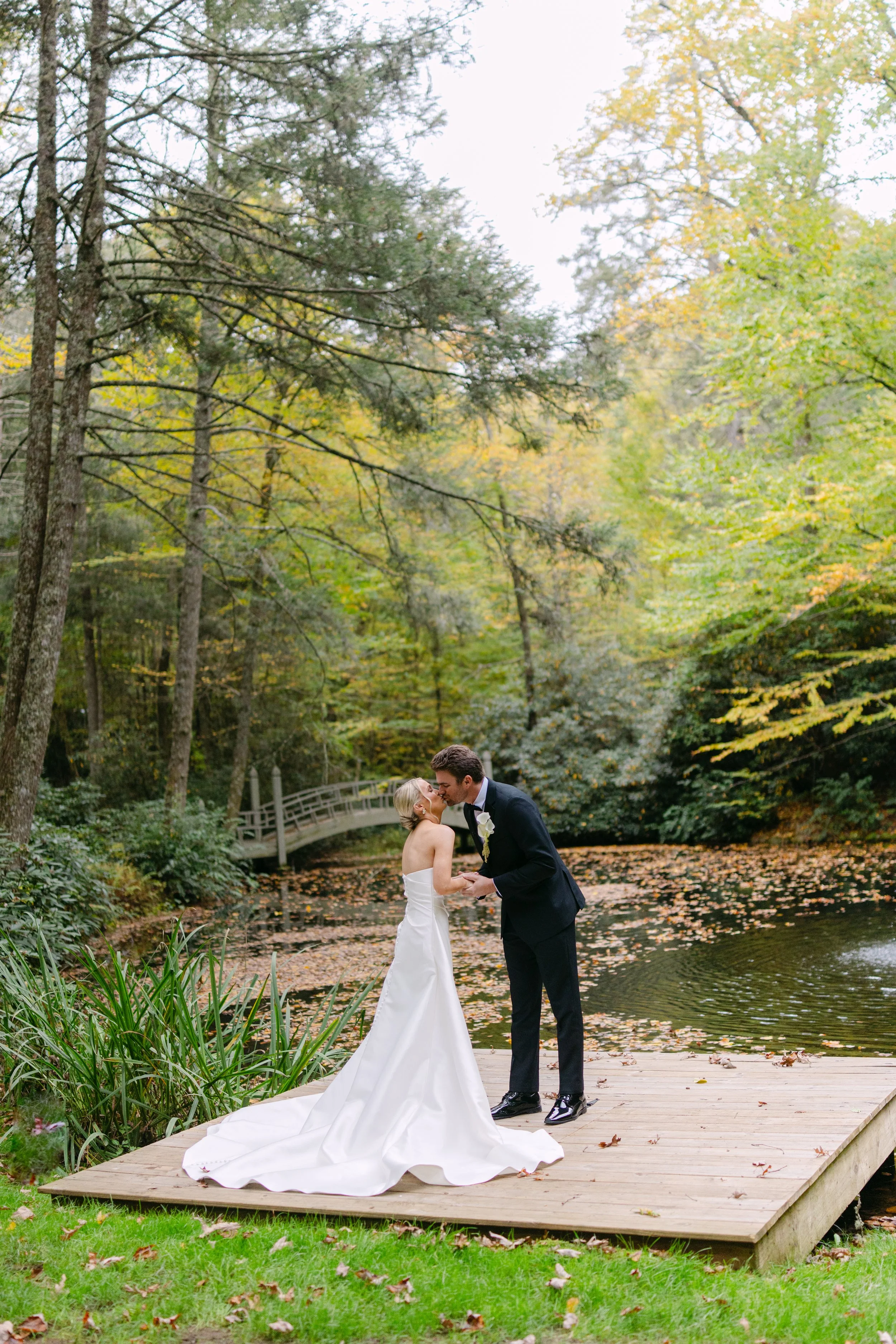 Bride and groom kissing on a wooden bridge beside a pond in an autumn forest.