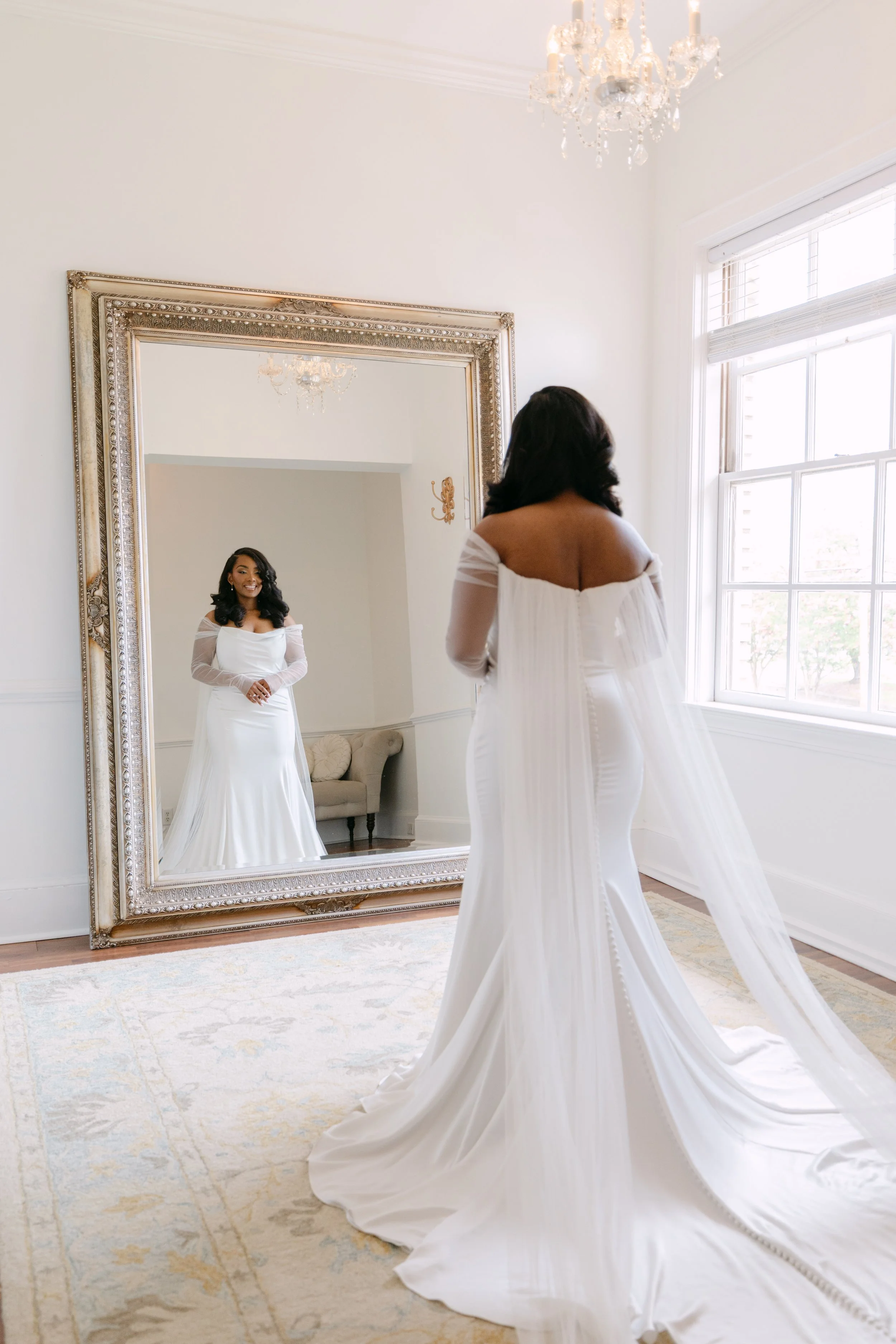 A woman in a white wedding dress is looking at her reflection in a large ornate mirror. She is inside a bright room with large windows, white walls, a chandelier, and a beige and white patterned rug.