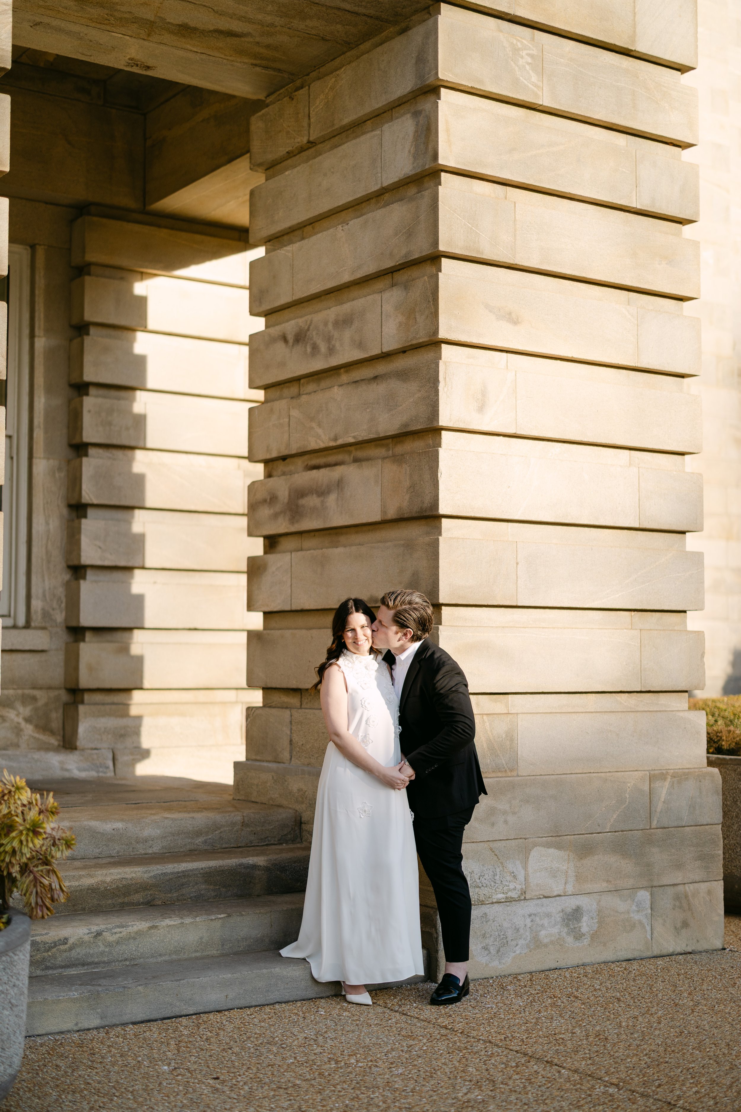 A couple in wedding attire standing outdoors against a large stone building, sharing a kiss.