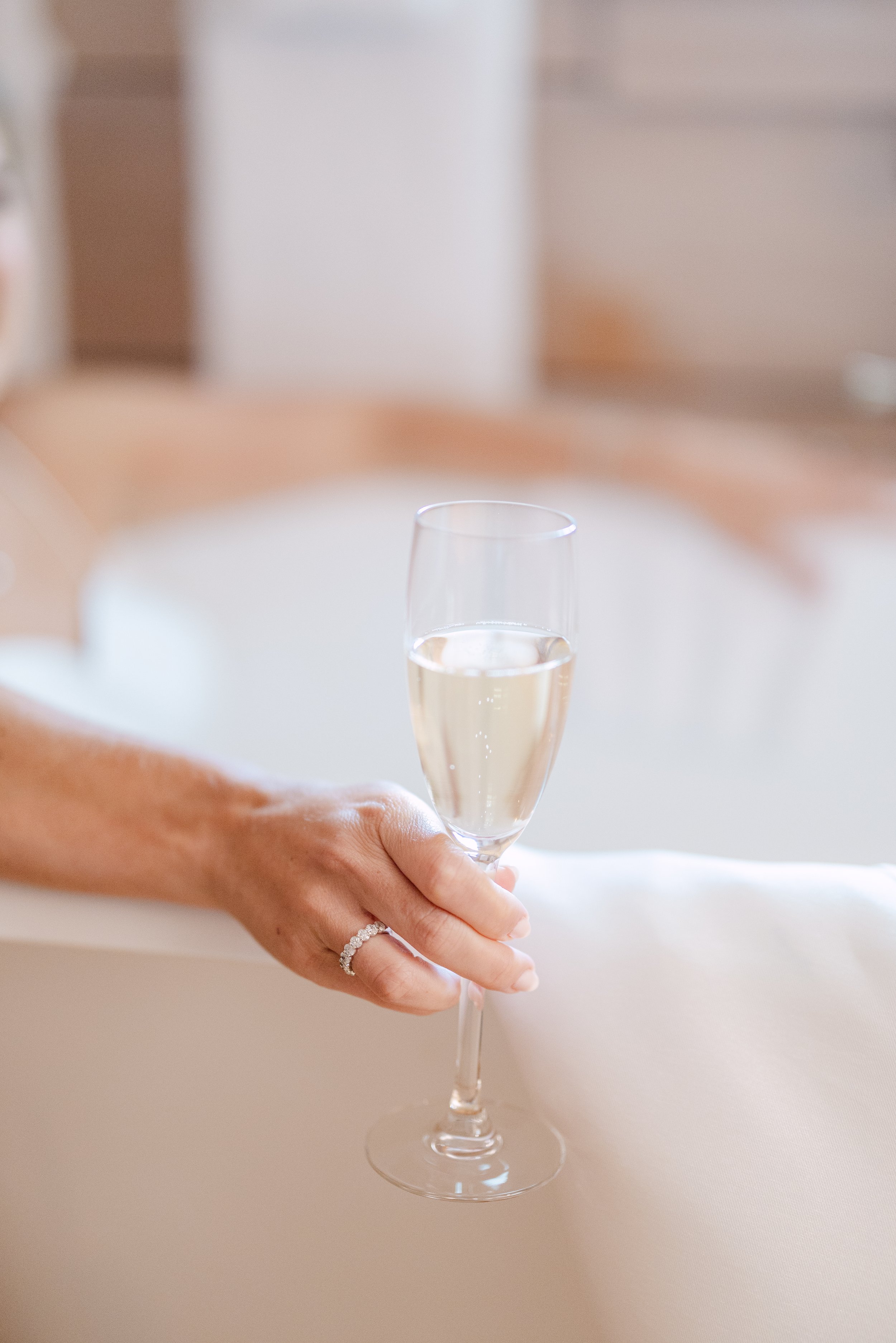 A person's hand holding a glass of champagne with an engagement ring on the finger, in a bright room with soft focus background.