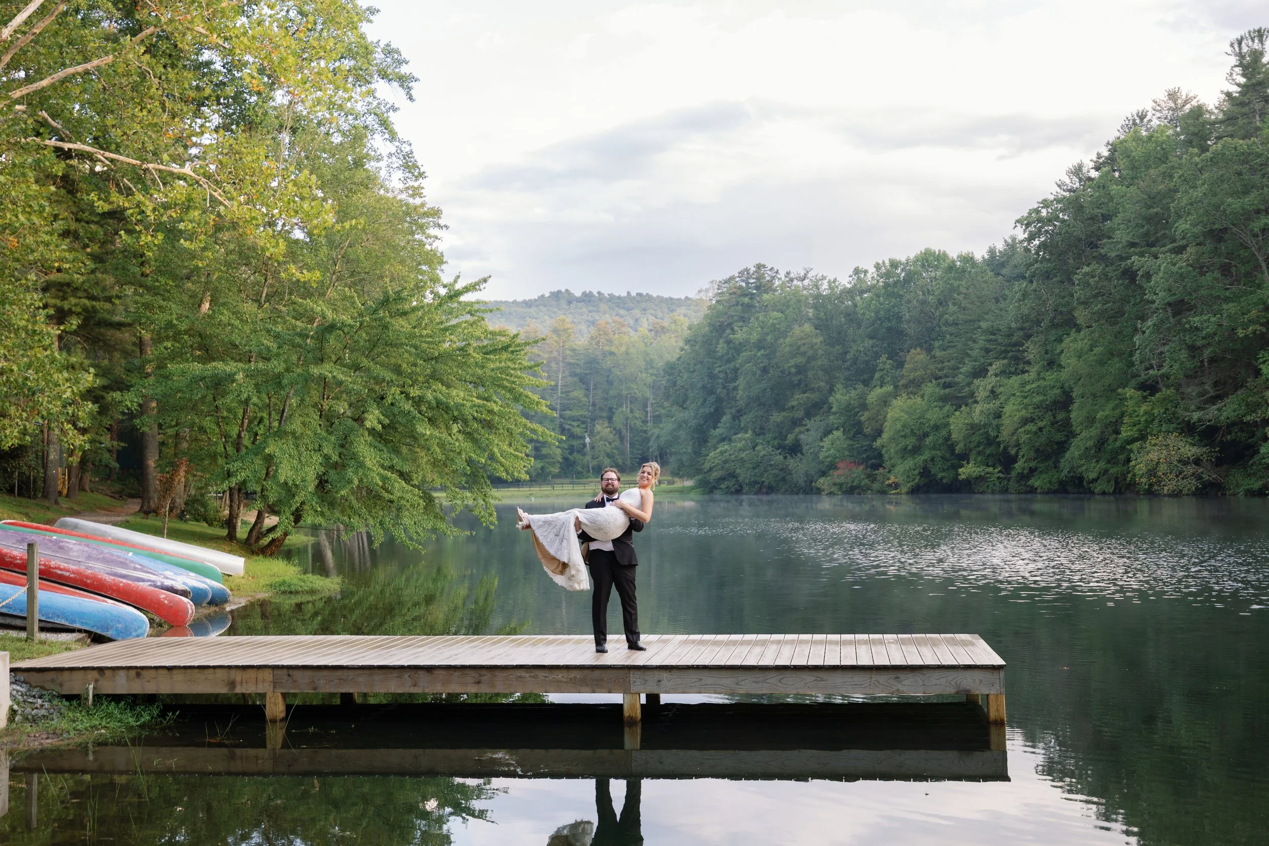 A wedding couple stands on a wooden dock by a lake, with a man holding a woman in a white wedding dress. Canoes are stacked on the grassy shore to the left, and lush green trees line the lake in the background, with hills and clouds in the sky.