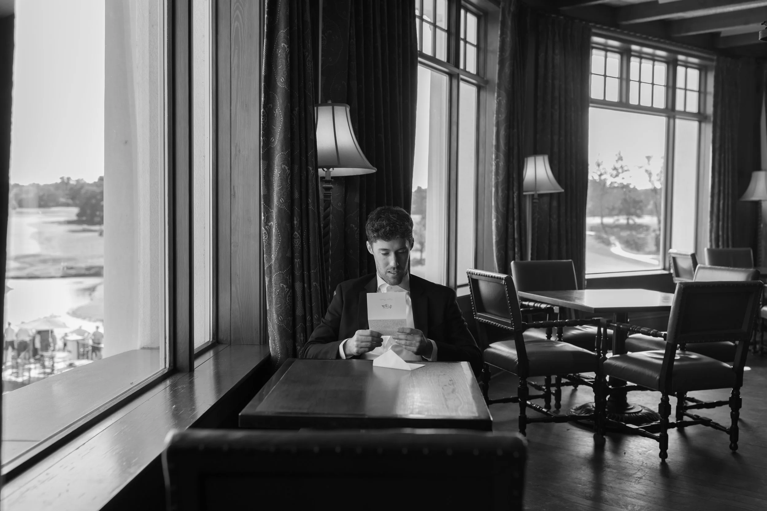 A man in a suit sitting alone at a table in a restaurant or cafe, reading a piece of paper, with large windows and curtains behind him.