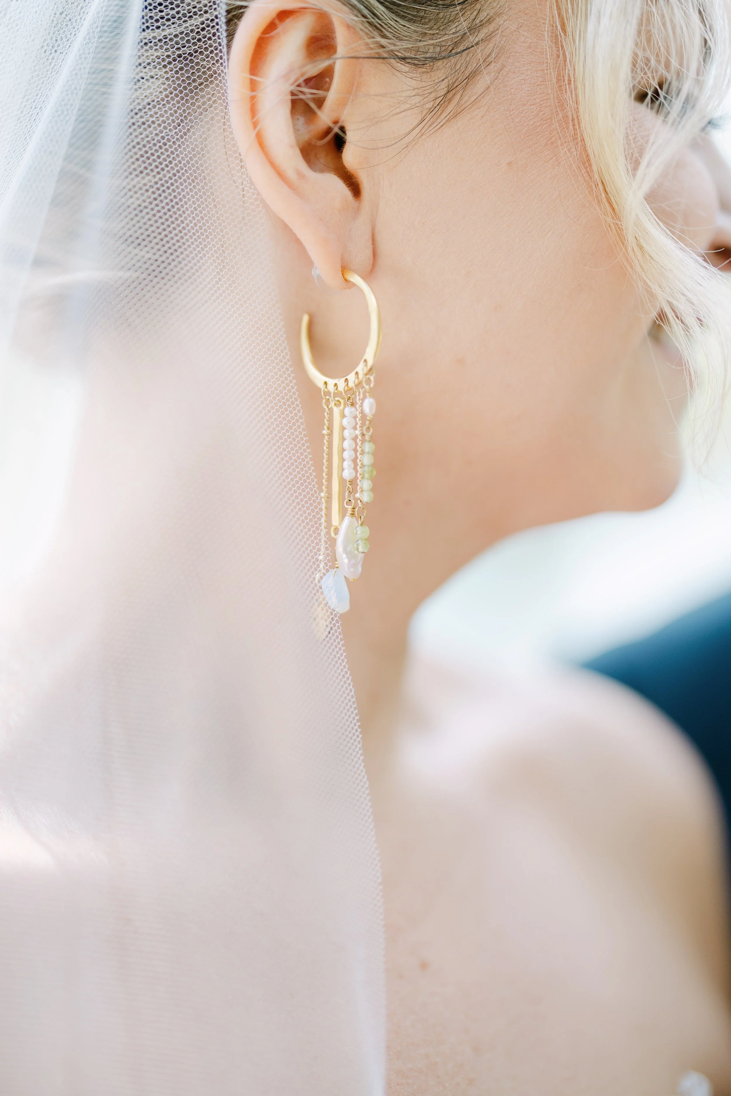 Close-up of a woman with blonde hair wearing a gold hoop earring with hanging pearl and chain details, and a sheer veil over her face.