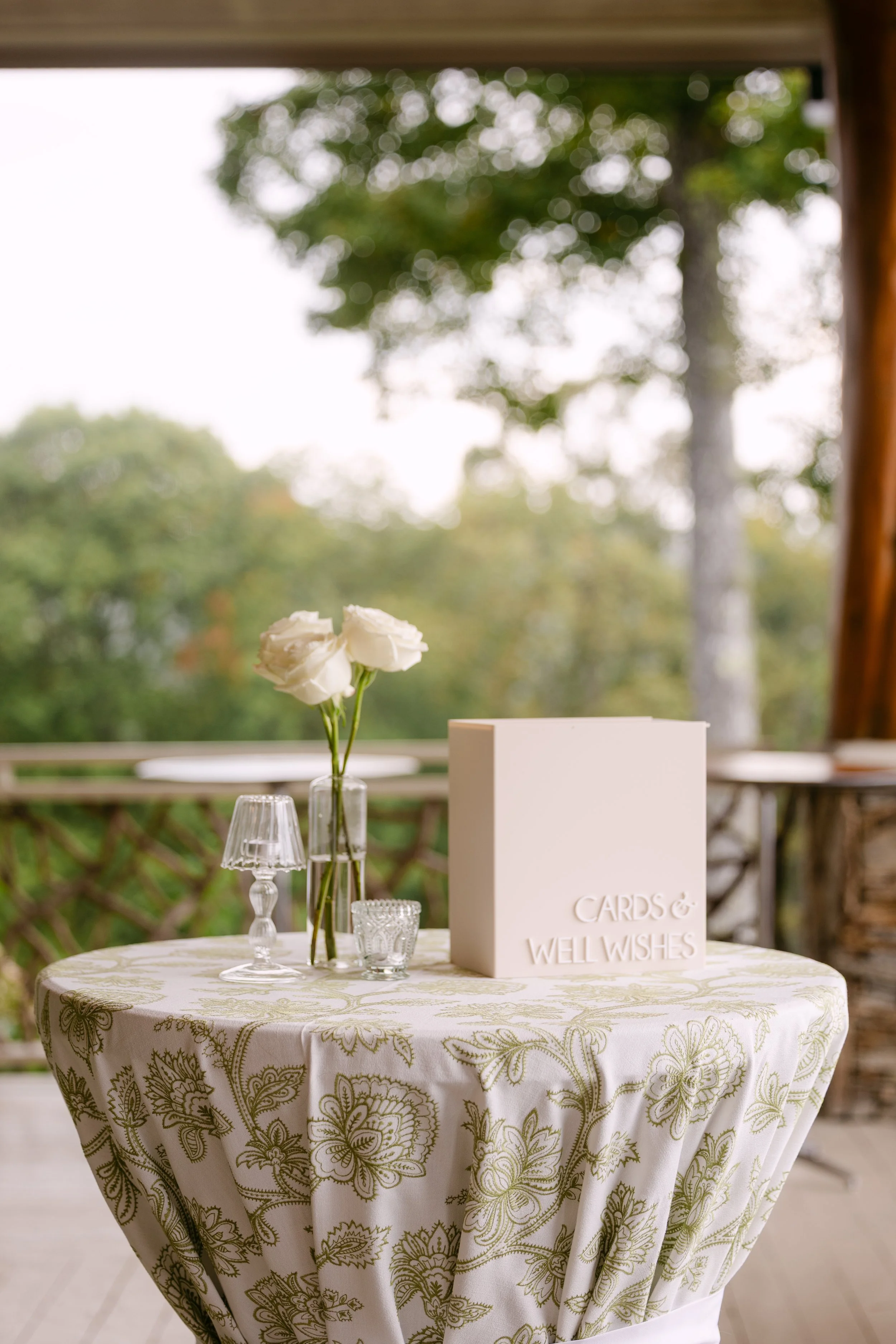 Table decorated with a white floral tablecloth, a small flower arrangement, a card box labeled "Cards & Well Wishes", and small decorative glass items, set outdoors with trees in the background.