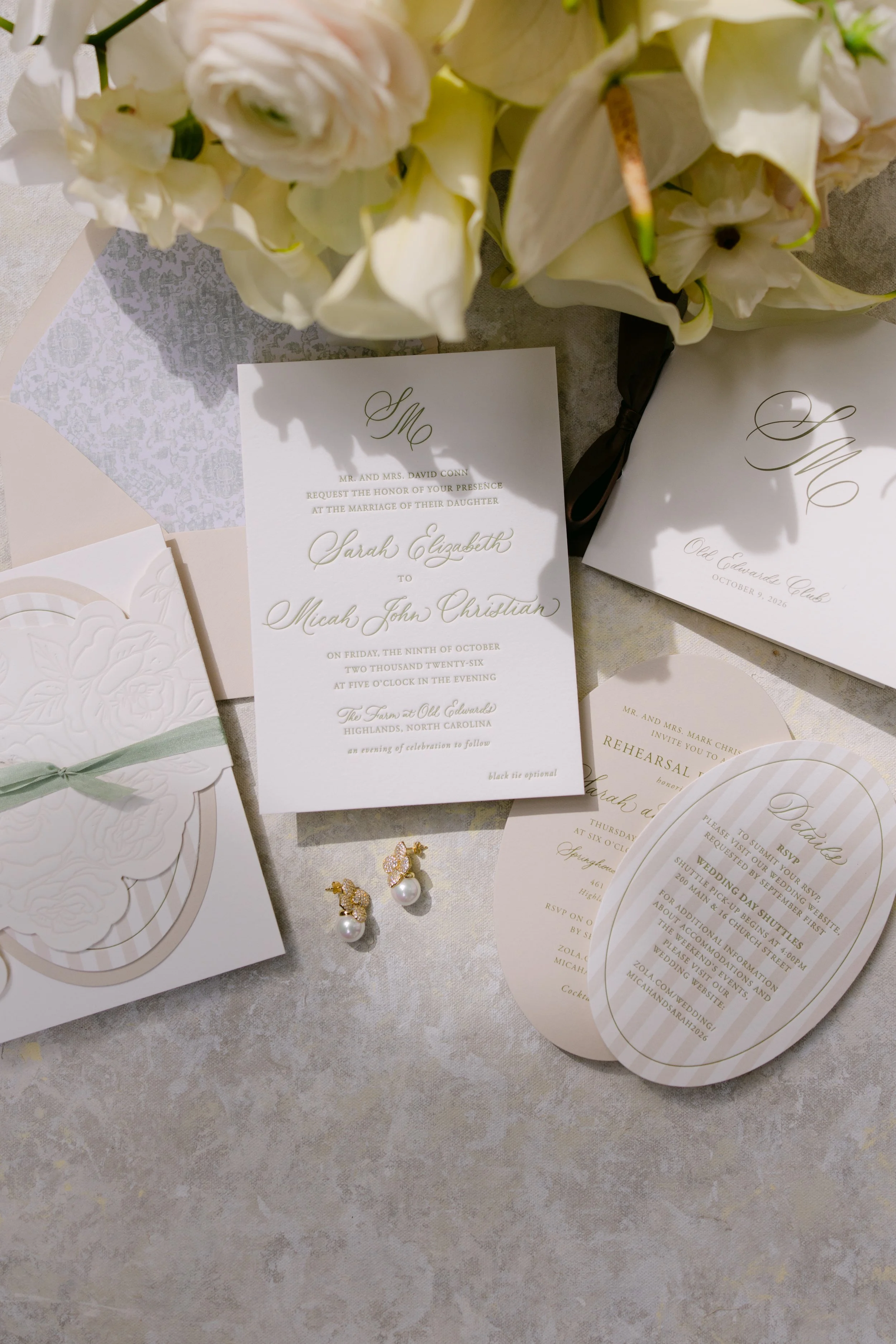 Flat lay of elegant white wedding invitations, floral arrangements, embossed stationery, pearl earrings, and event details.