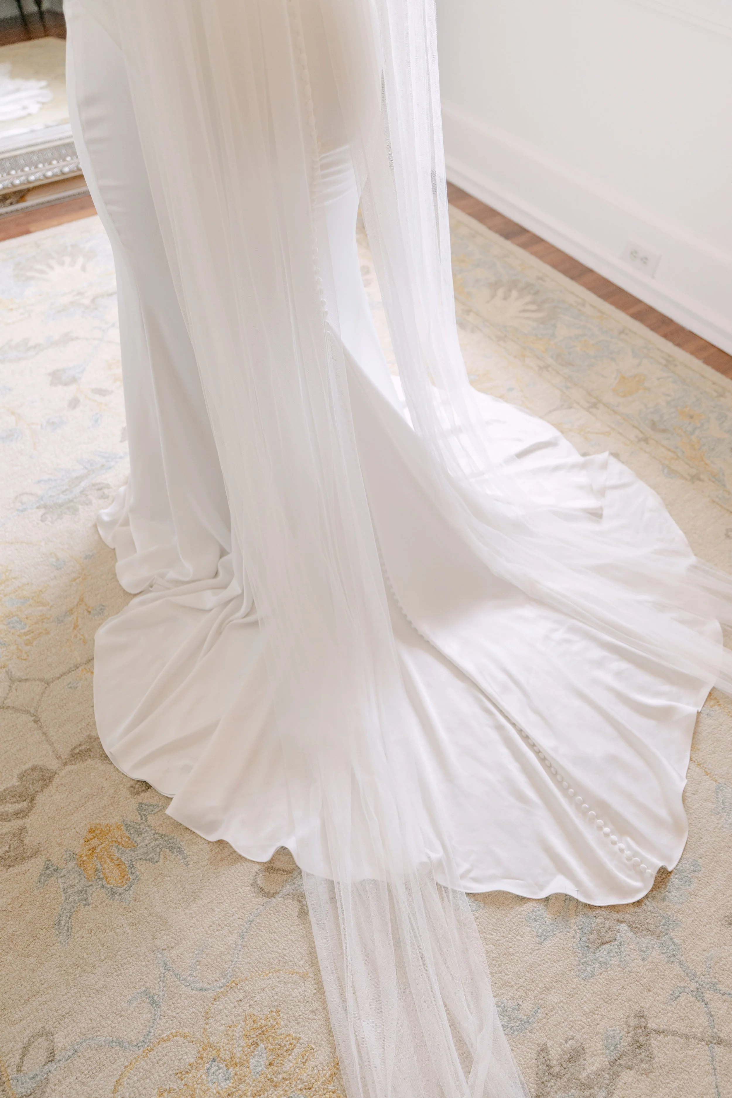 Close-up of a white wedding dress with a long train, hanging in a room with a light-colored patterned rug and a mirror in the background.