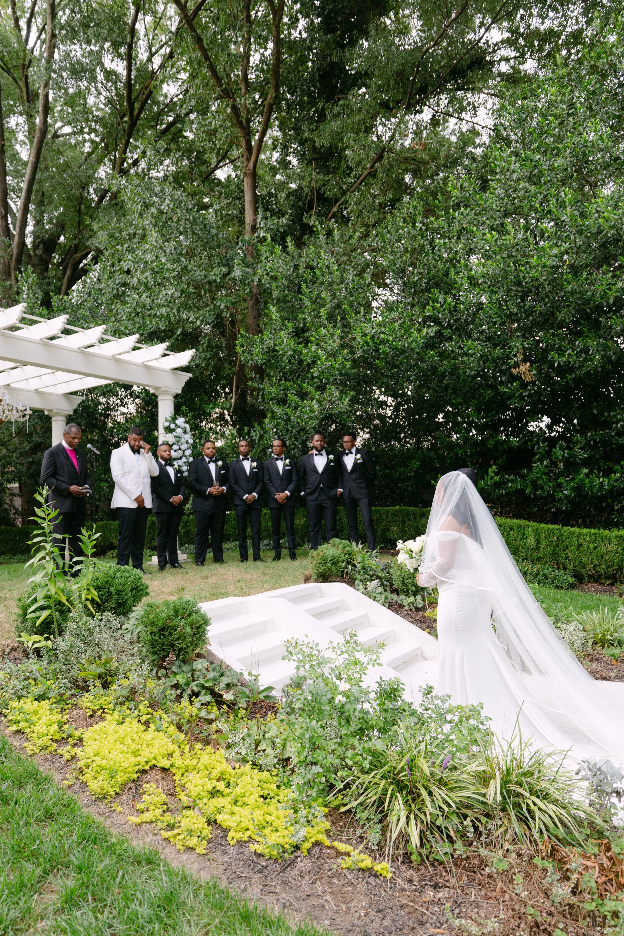 An outdoor wedding ceremony with a bride in a white dress and veil kneeling in prayer or reflection, facing a group of seven groomsmen and one officiant, surrounded by lush green trees and plants, with white flowers and a decorative white structure o