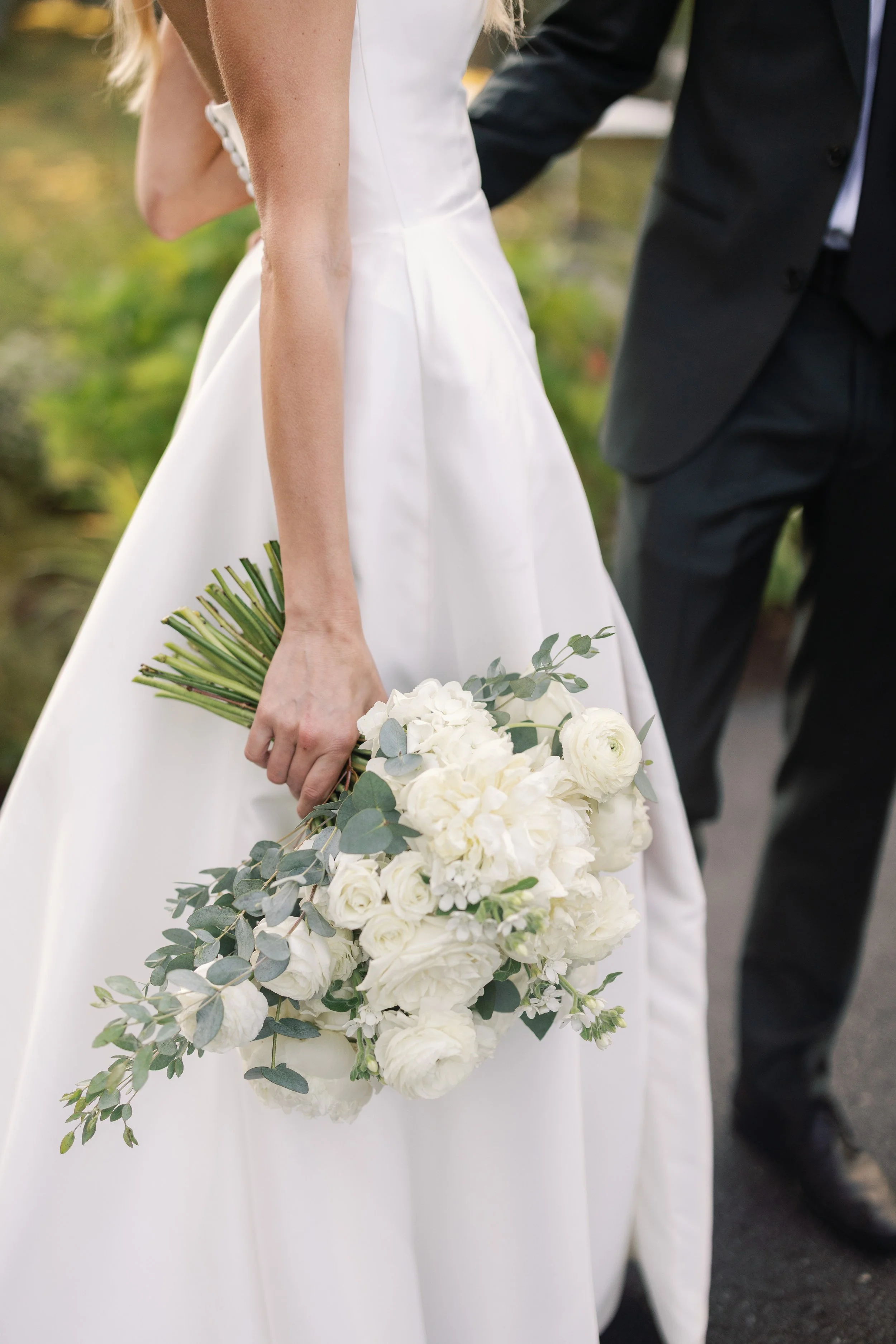Bride holding a bouquet of white flowers and greenery, standing next to a groom dressed in a black suit.