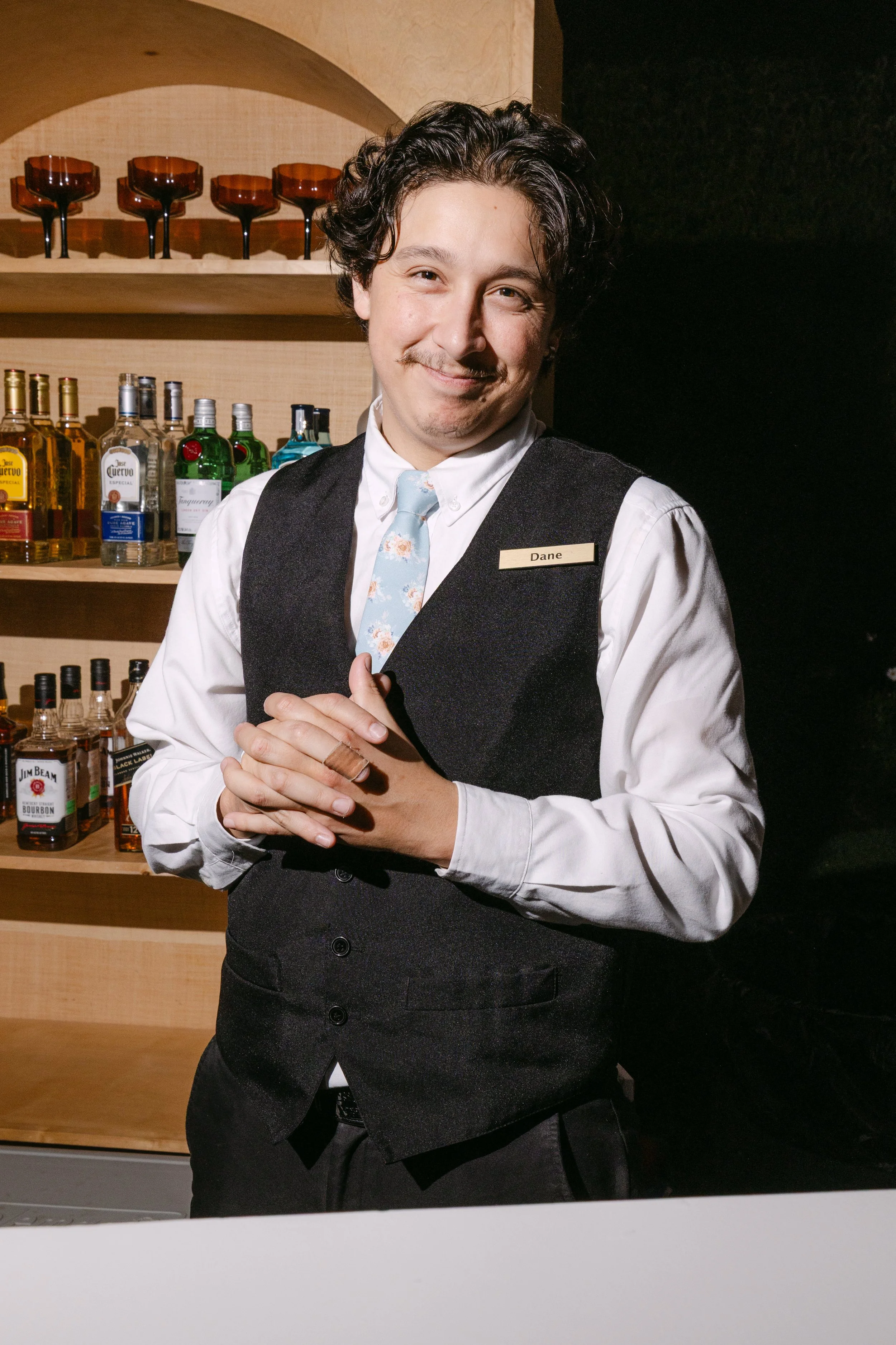 A man with a name tag that reads 'Dane' standing behind a bar with various bottles of alcohol in the background.