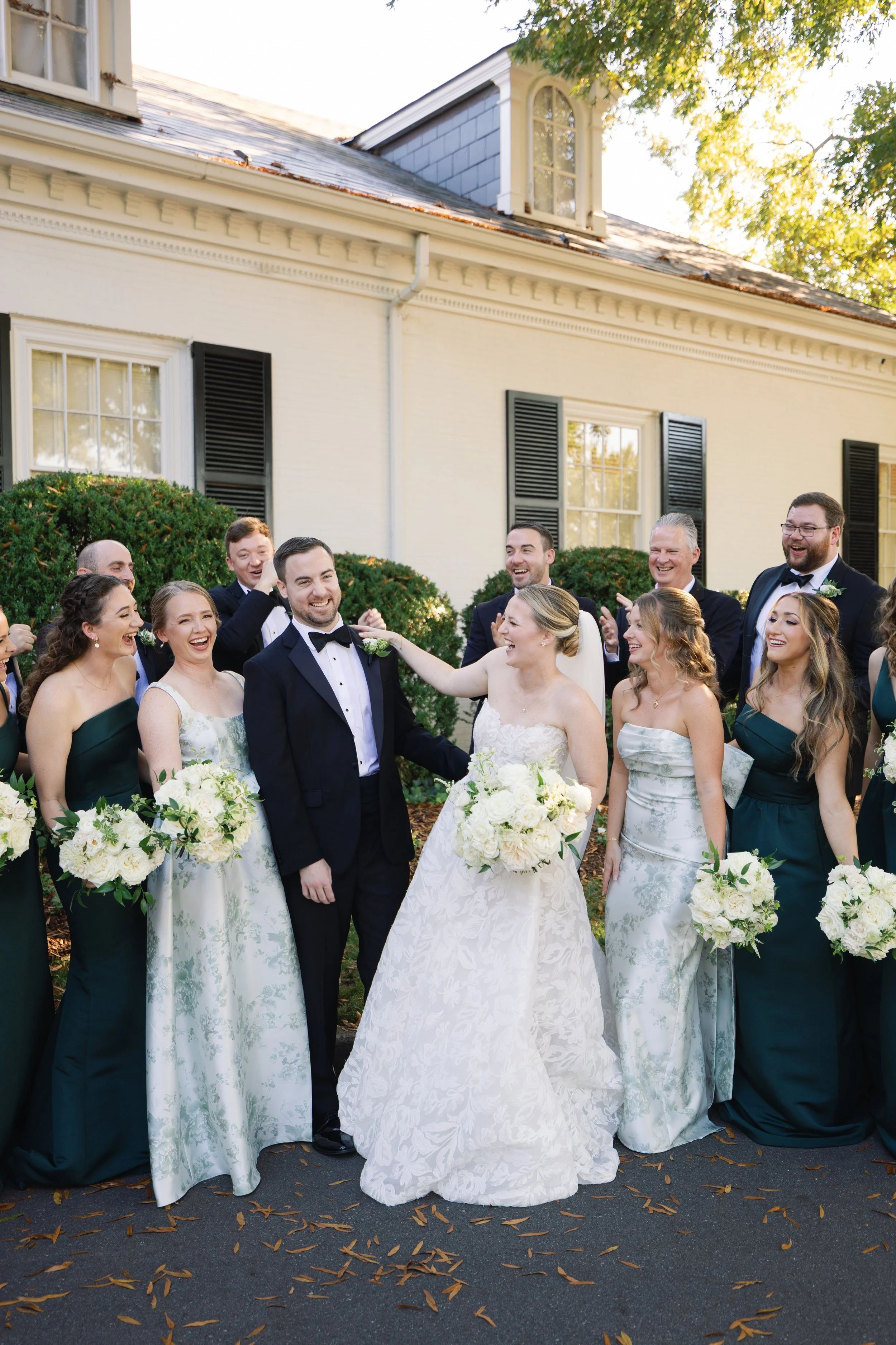 A group of people dressed in wedding attire celebrating outside a house with greenery and trees.
