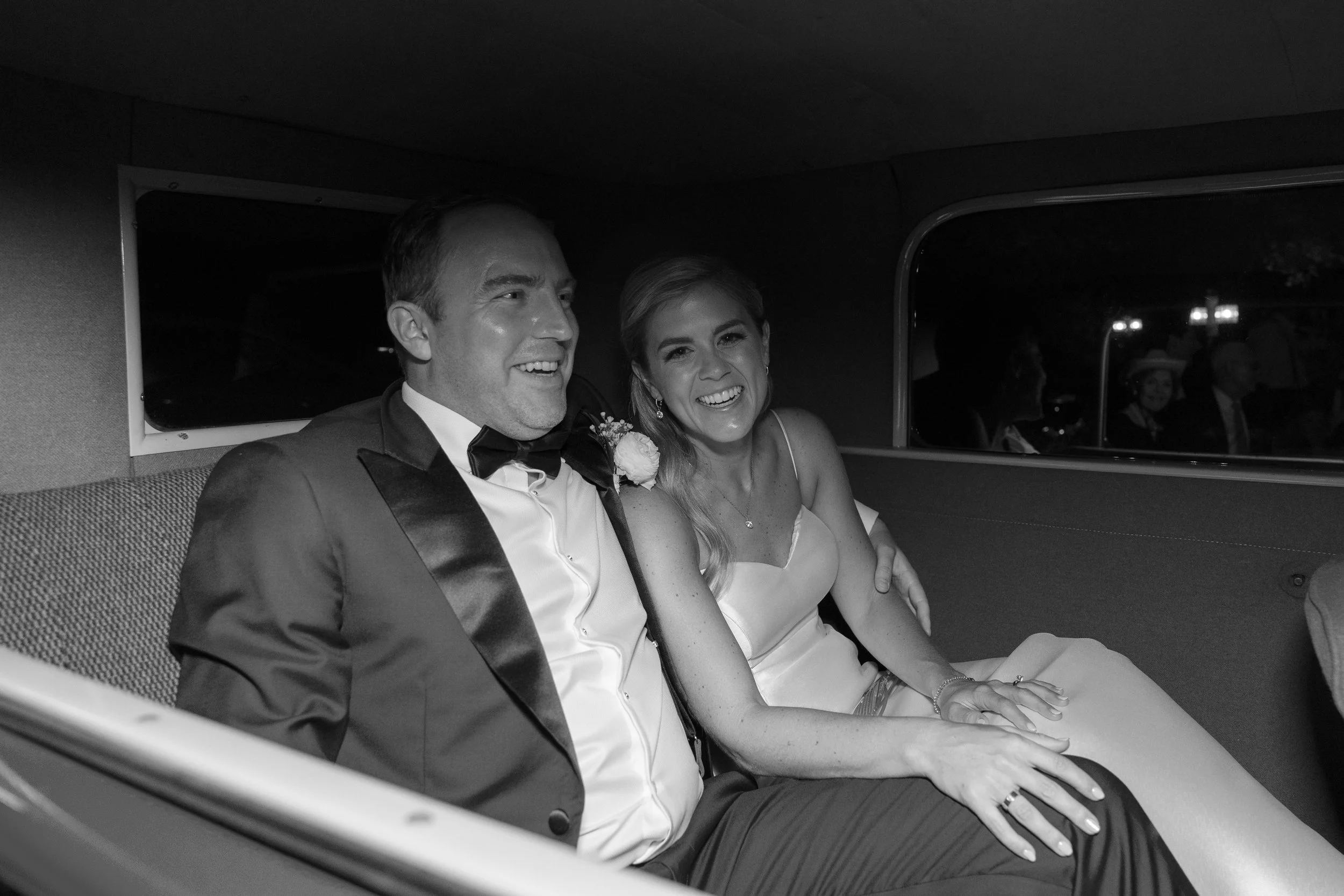 A black and white photo of a smiling couple dressed in wedding attire sitting inside a limousine. The groom is in a tuxedo, and the bride is in a wedding dress with a ring on her finger.