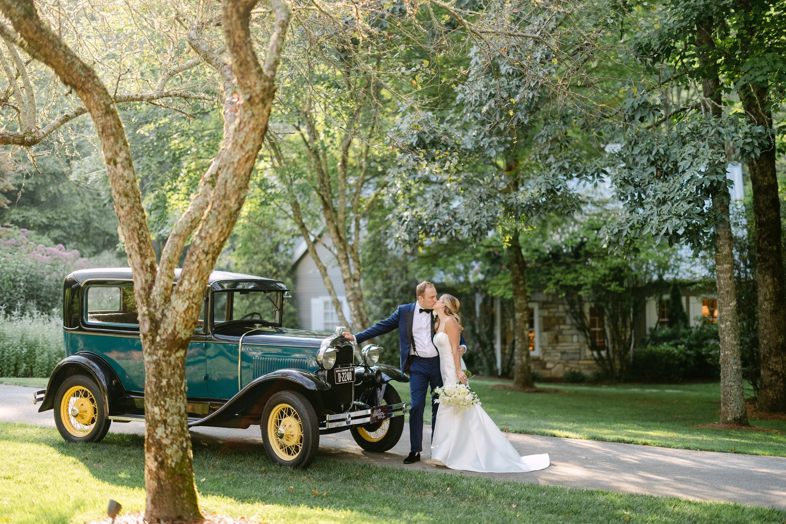 A bride and groom kissing beside a vintage black and teal car on a driveway surrounded by green trees and a house in the background.