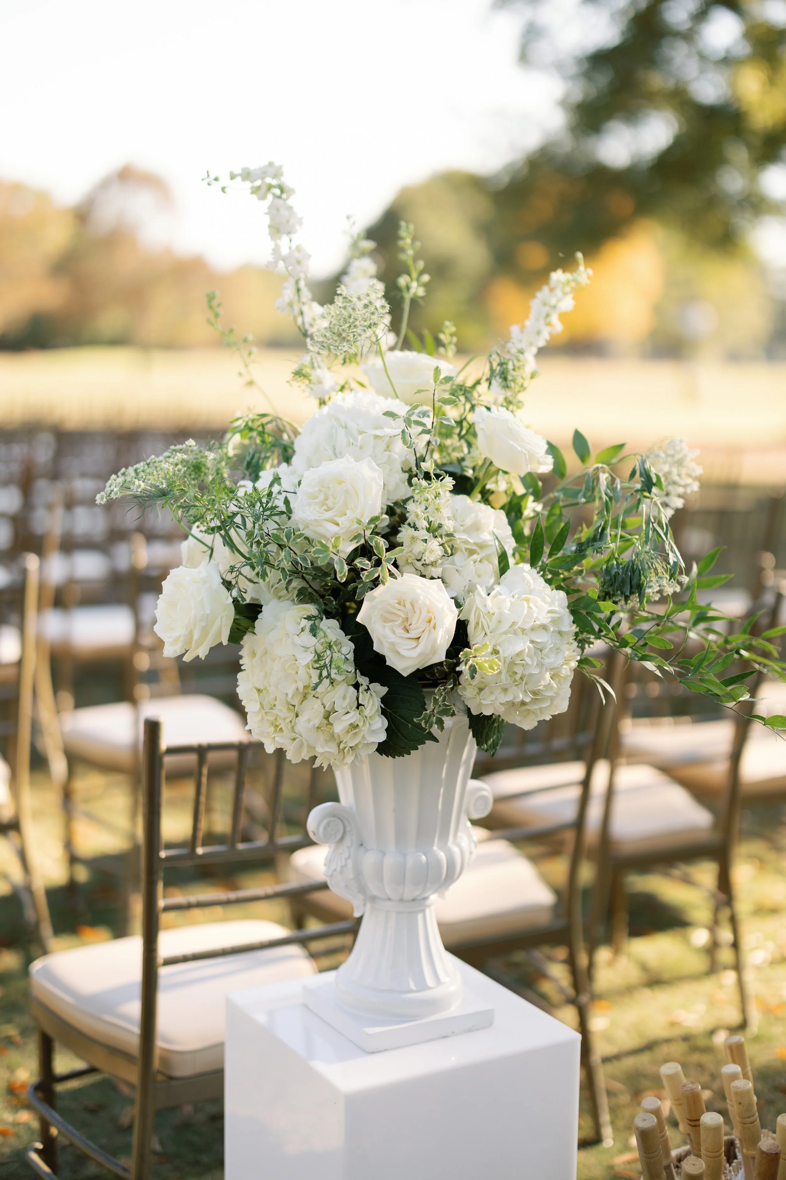White floral arrangement with roses and hydrangeas in a tall white vase at an outdoor event.