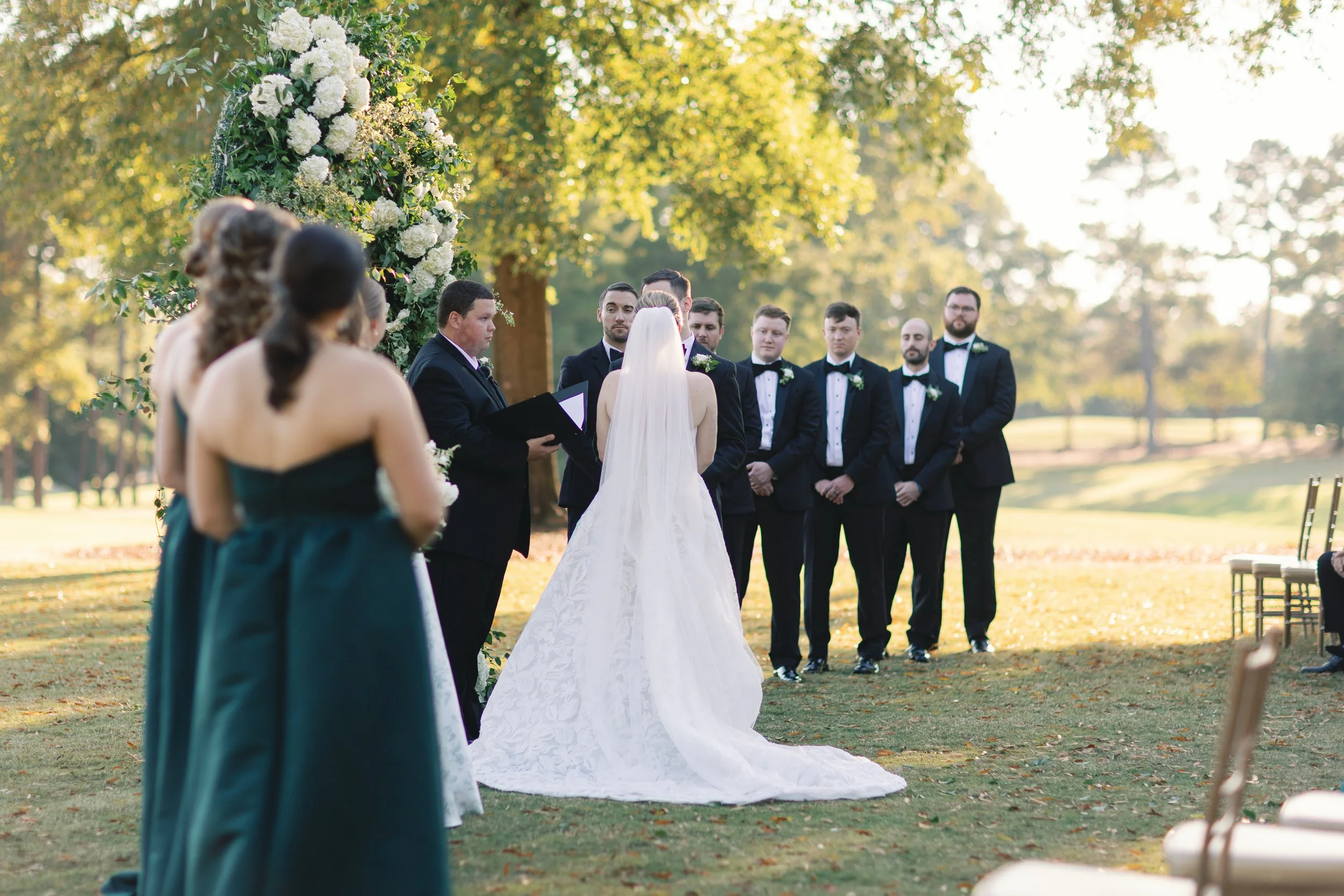 A bride in a white wedding gown with a long train and veil standing under a floral arch, facing a wedding officiant and groom, with bridesmaids and groomsmen in formal attire on either side, outdoors on a sunny day with trees in the background.