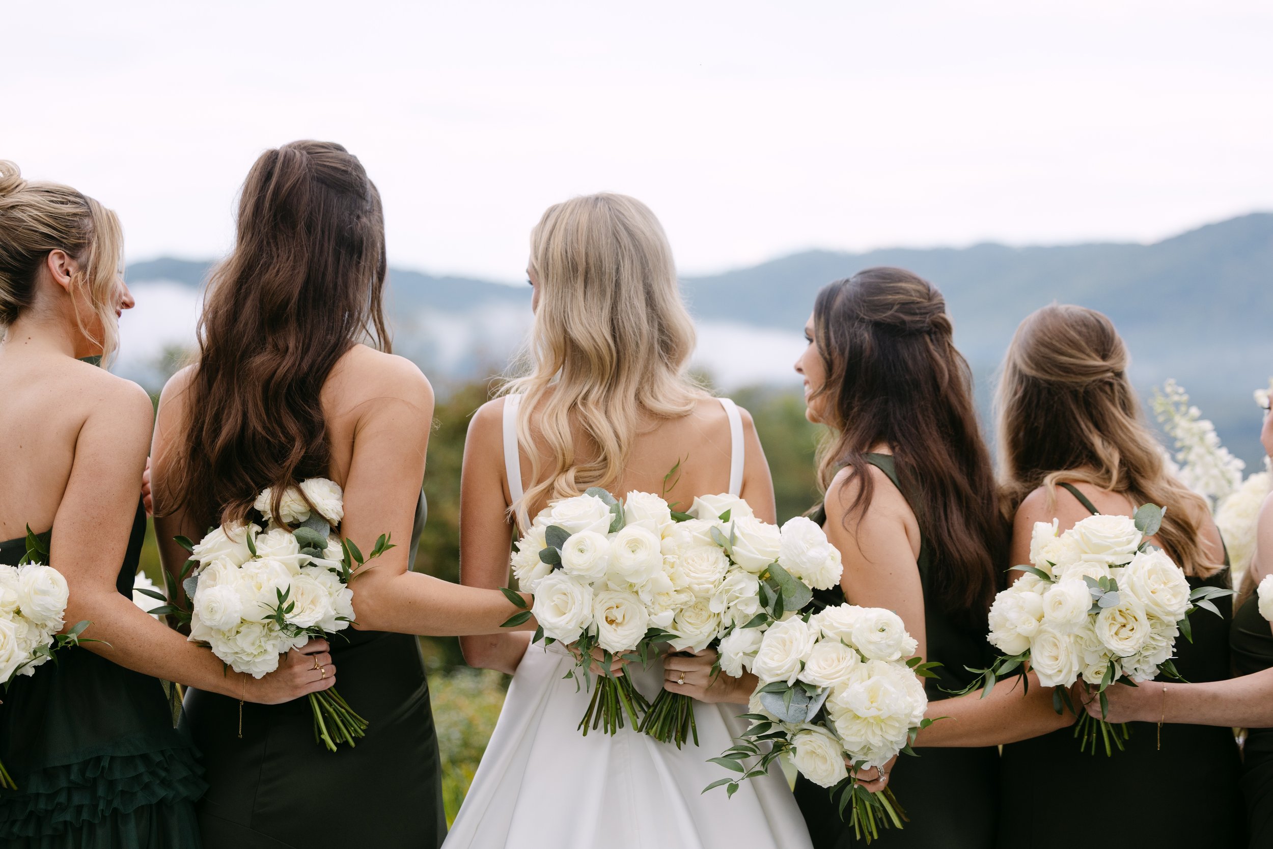 Group of women in bridesmaid and bride dresses holding white bouquets, standing outdoors with mountains in the background.