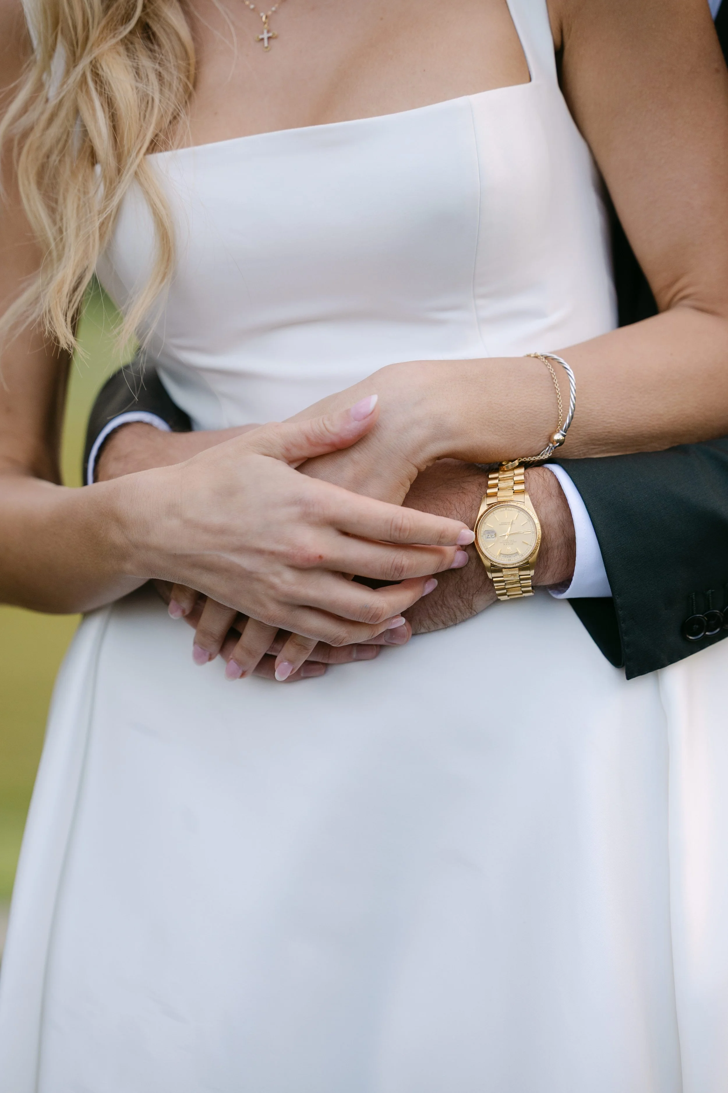 Close-up of a woman in a white dress and a man in a suit embracing, with focus on their hands intertwined, showing a gold watch and jewelry.