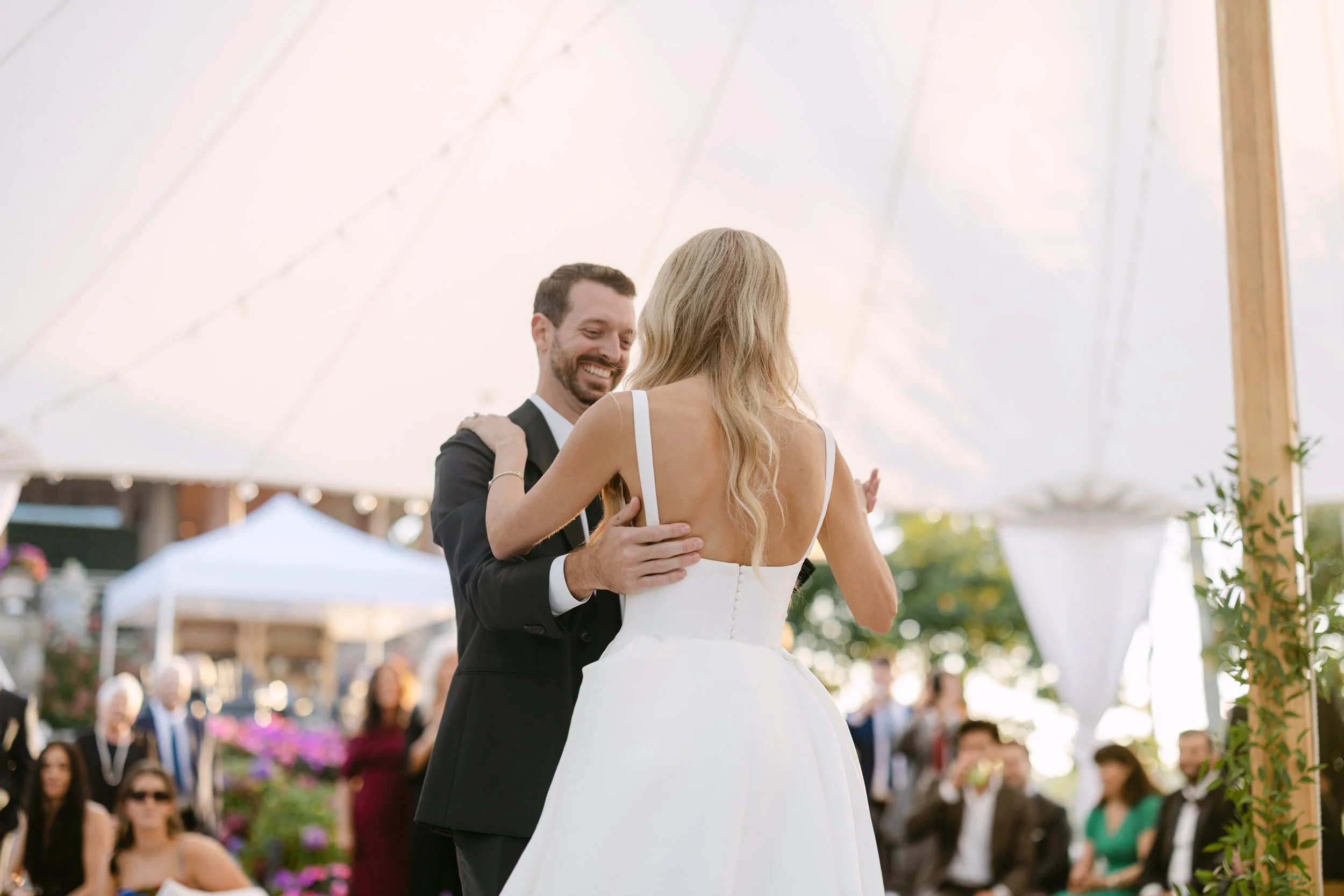 A couple dancing at their wedding reception under a white tent, with guests watching in the background.