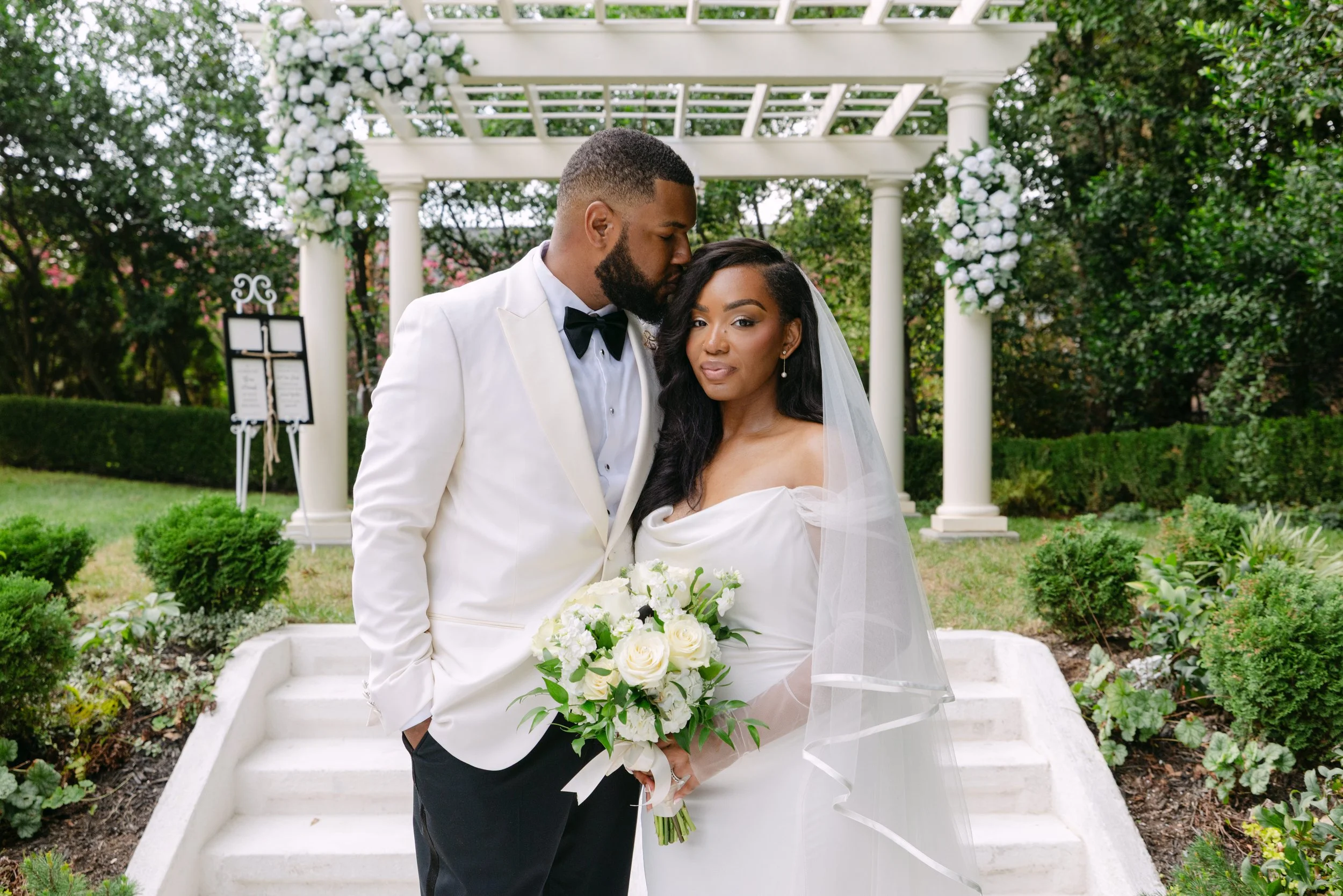 A bride and groom standing close together outdoors, with the groom kissing the bride's temple. The bride is holding a bouquet of white roses, wearing a white off-the-shoulder wedding dress with a veil. The groom is in a white tuxedo jacket with a bla