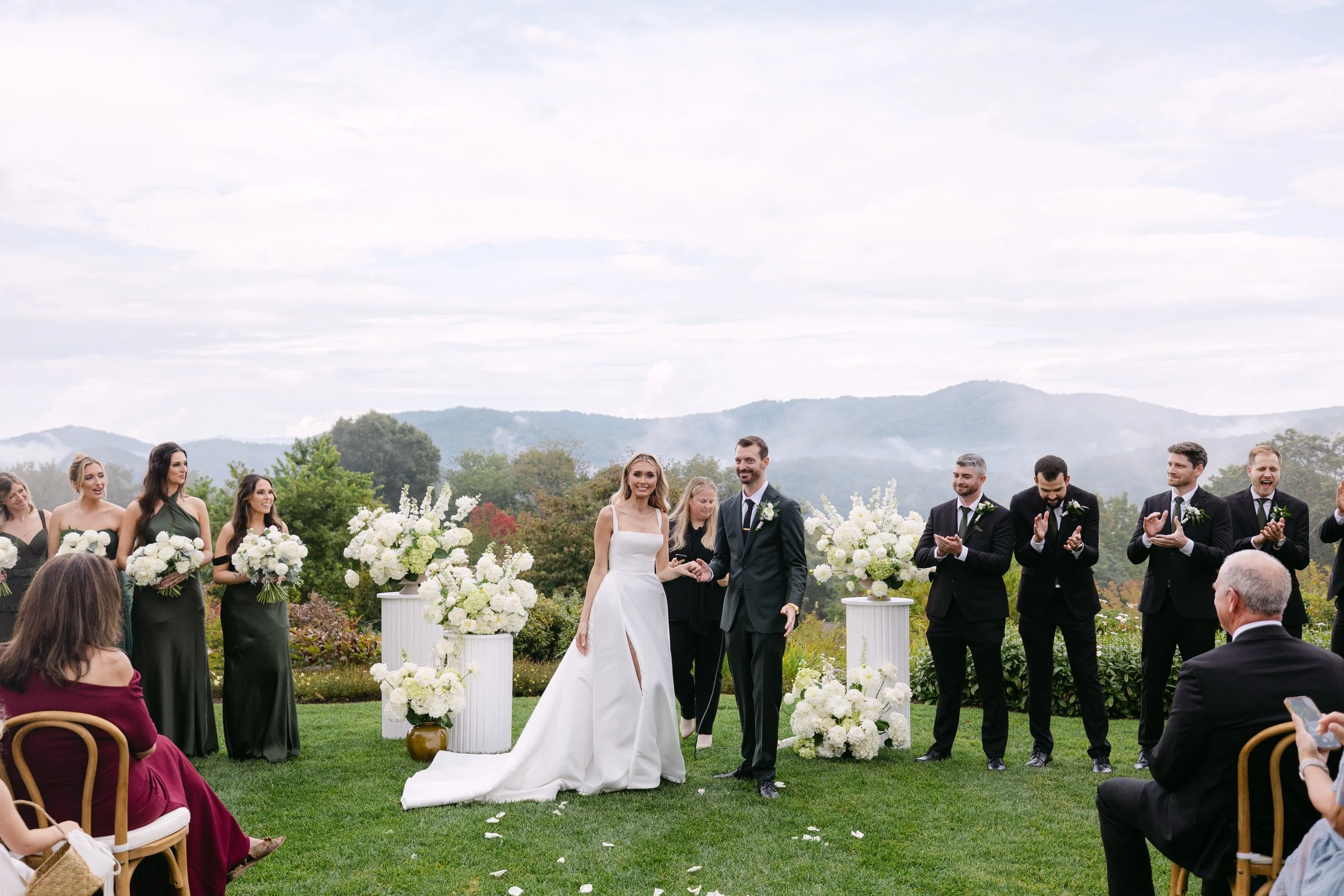 A wedding ceremony outdoors with the bride and groom walking down the aisle, surrounded by bridesmaids and groomsmen, with guests seated on either side.