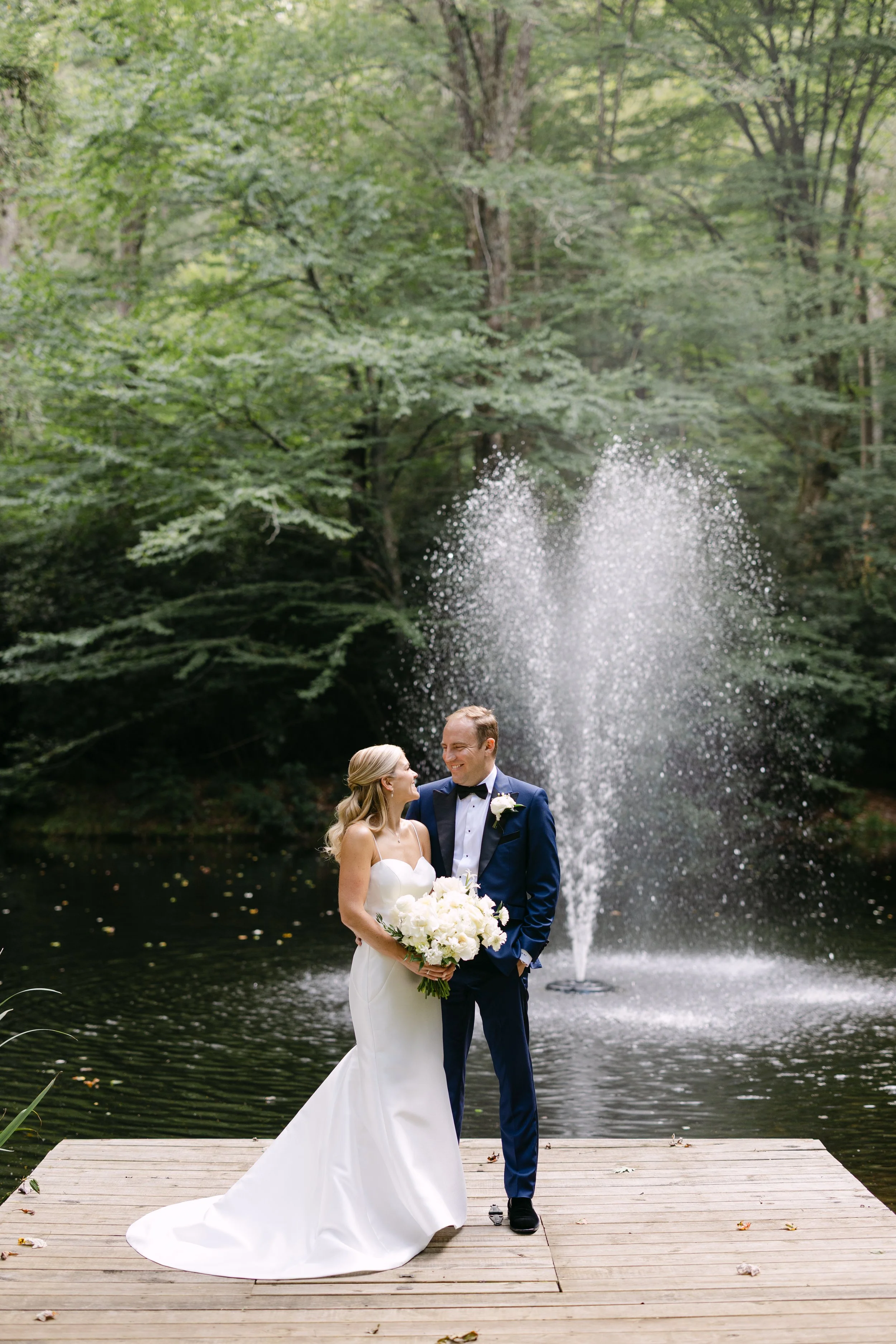 A couple in wedding attire standing on a wooden dock by a pond, with a fountain spraying water behind them, surrounded by green trees.