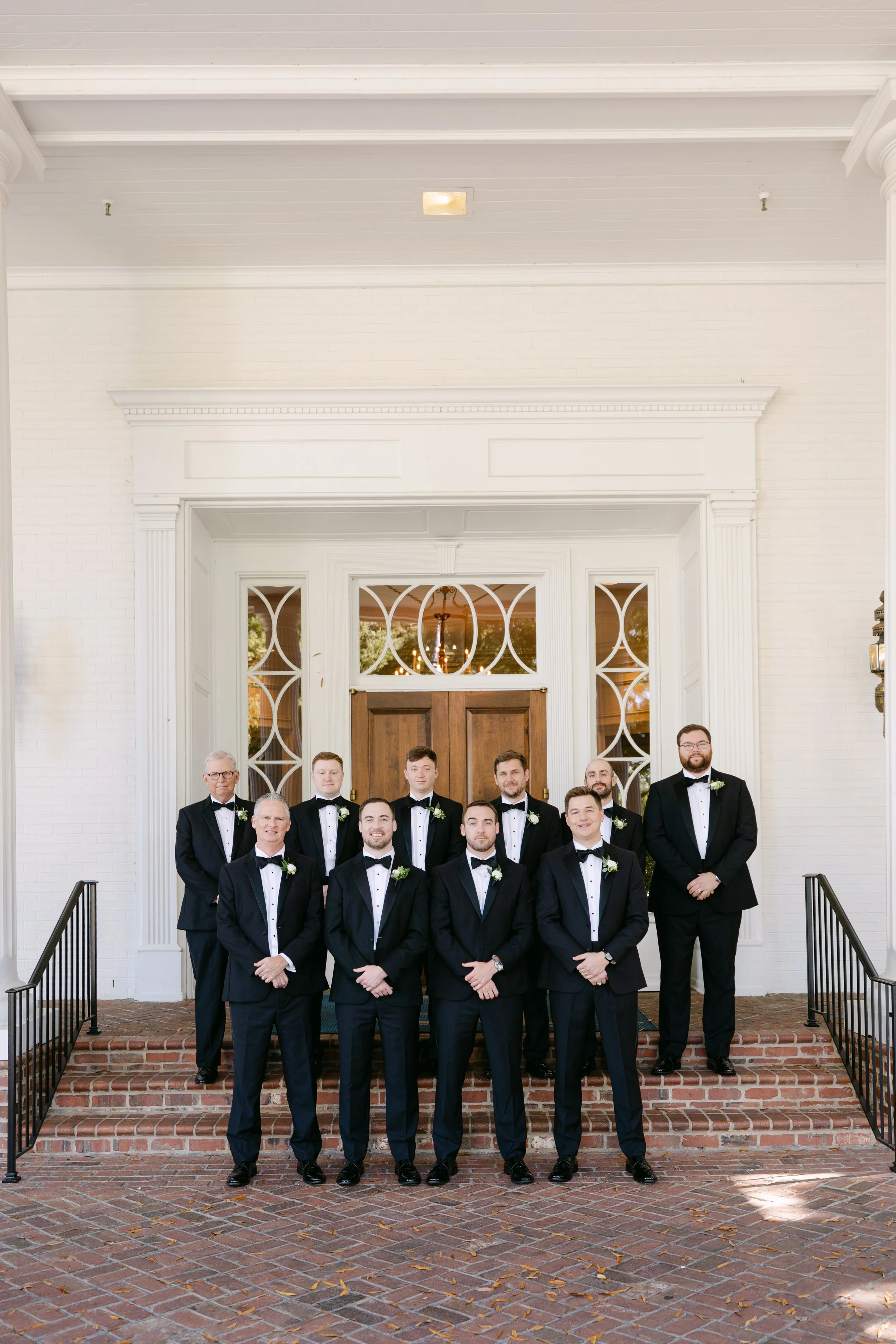 Group of men in tuxedos standing on brick steps in front of a white building with wooden door and decorative glass panels.