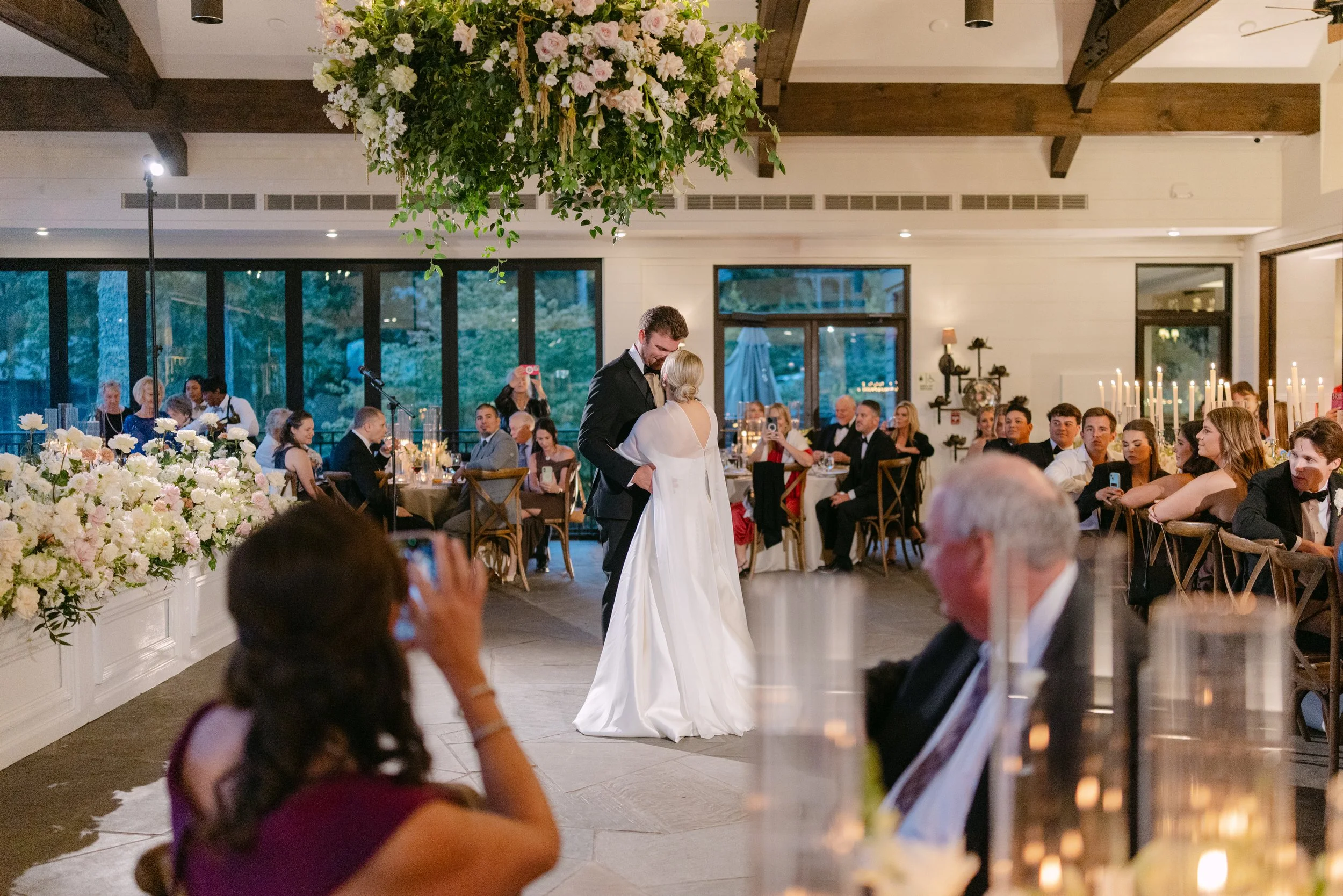 Bride and groom sharing a first dance at an elegant wedding reception surrounded by guests, candles, and floral arrangements