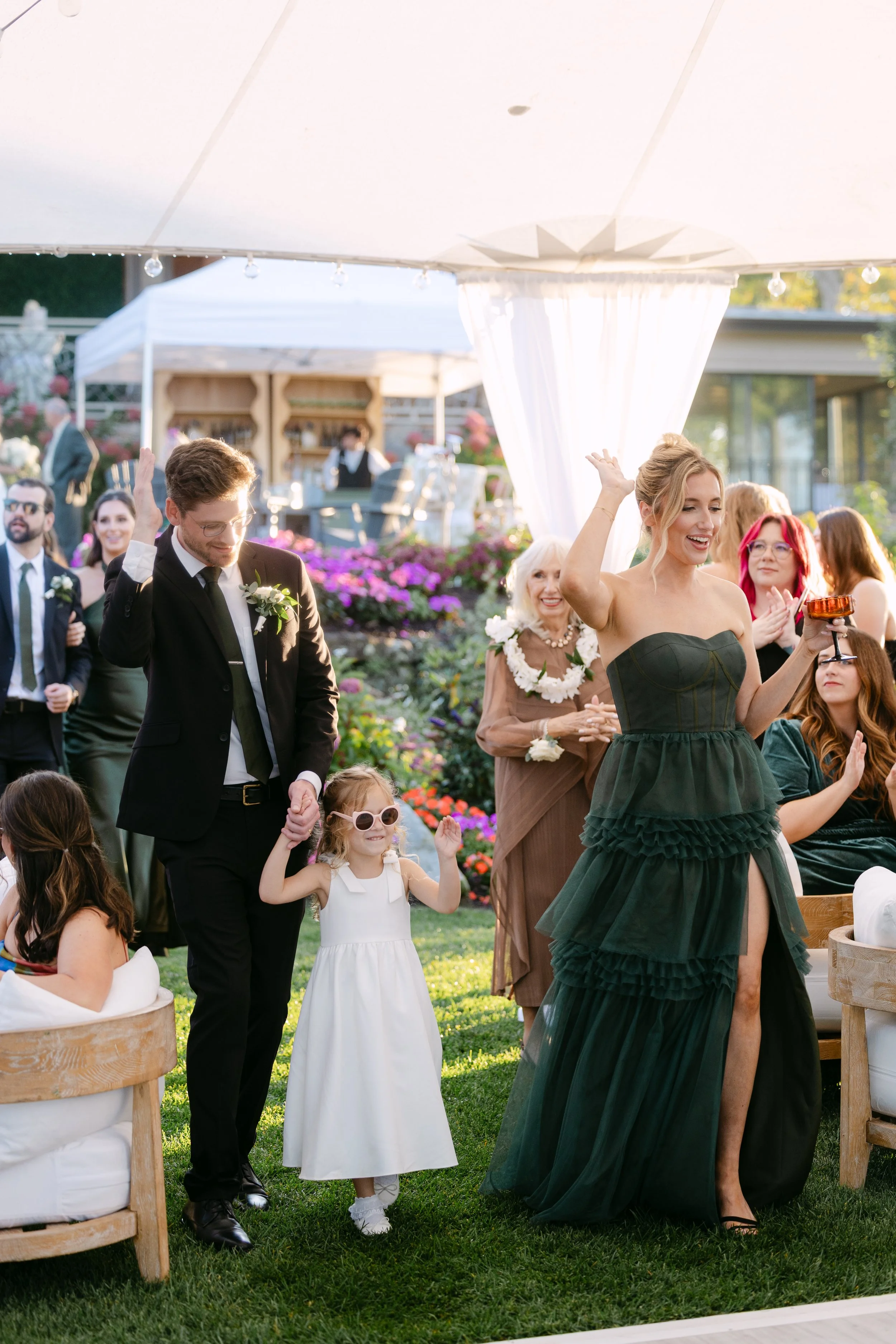 Group of people celebrating outdoors, including a young girl in sunglasses, a woman in a green strapless dress, and a man in a suit, at a garden event with flowers and a white canopy.