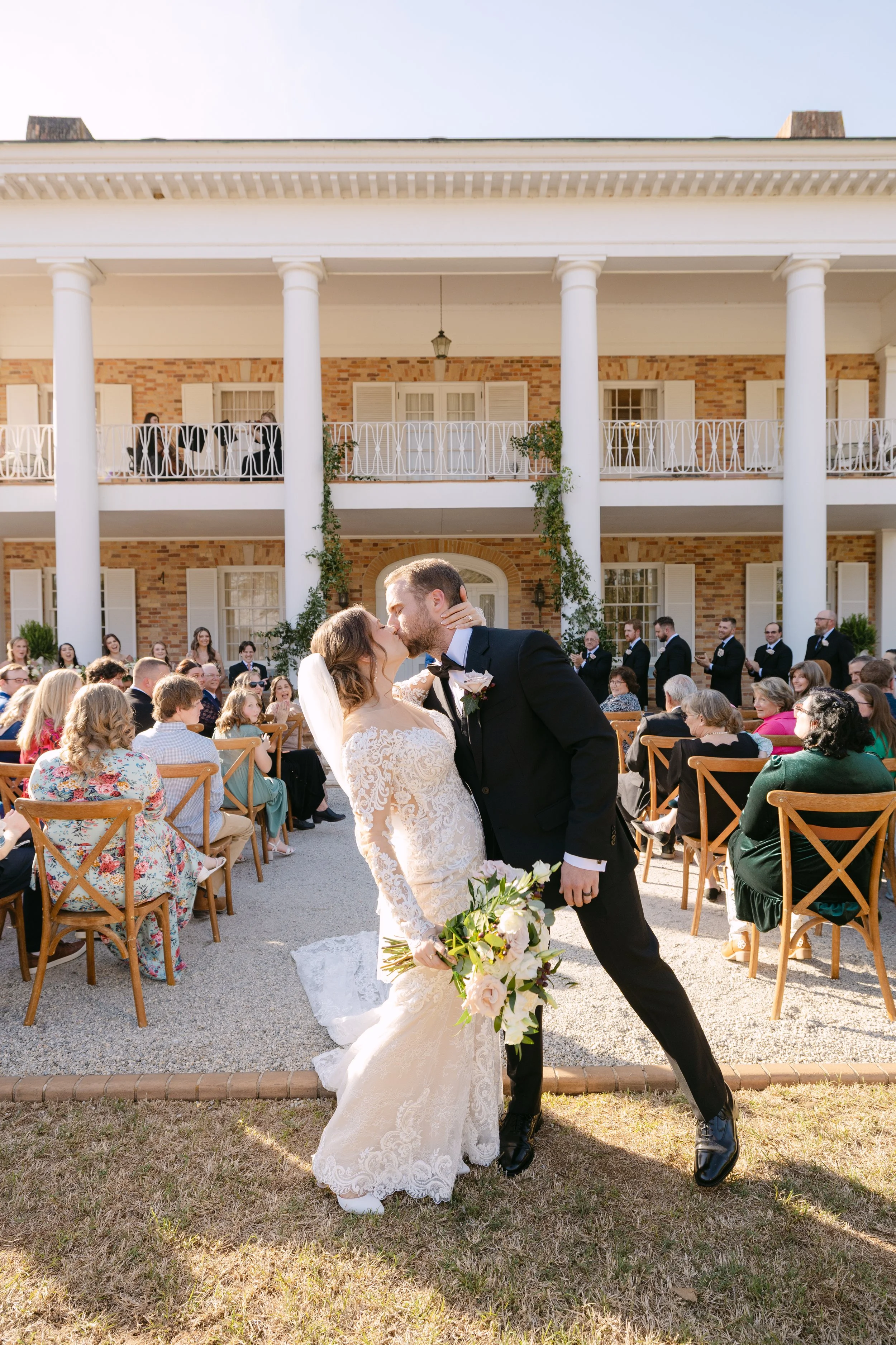 Bride and groom kissing at their outdoor wedding ceremony surrounded by seated guests, with a large brick and white-columned building in the background.