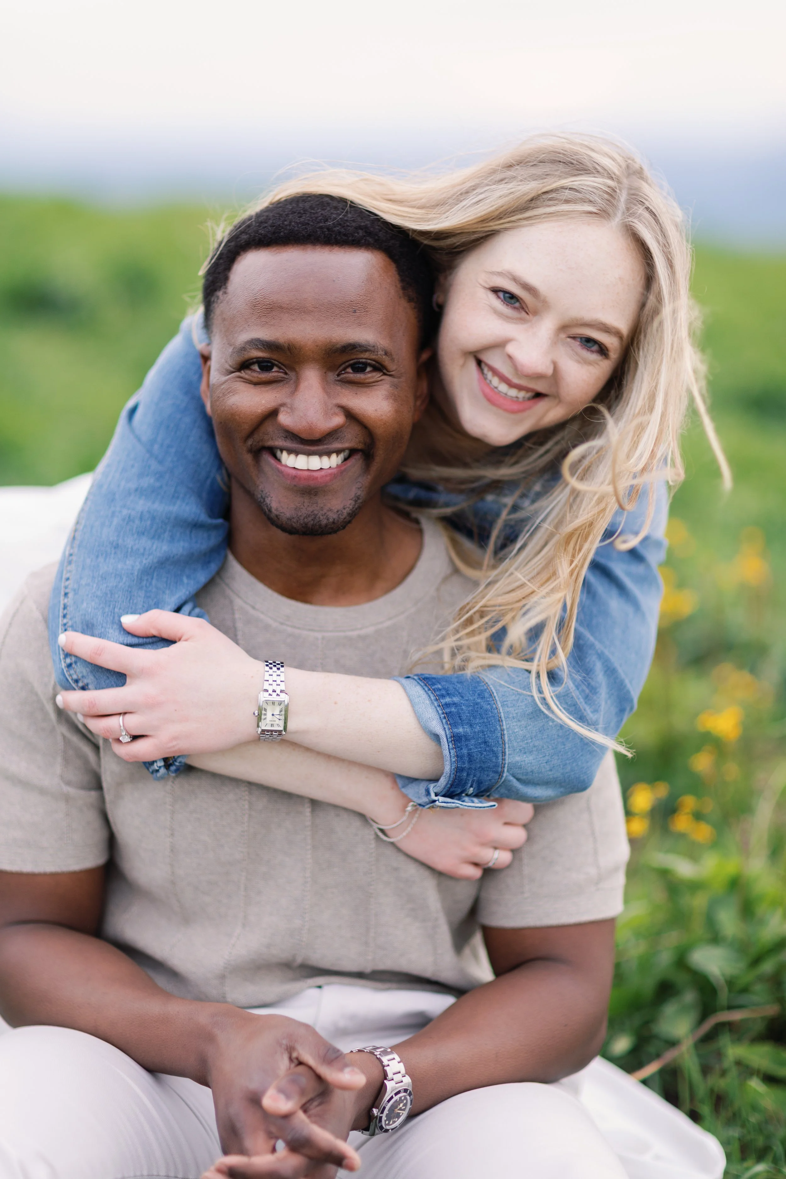 A smiling interracial couple outdoors, with the woman on the man’s back, in a green field, both looking at the camera.