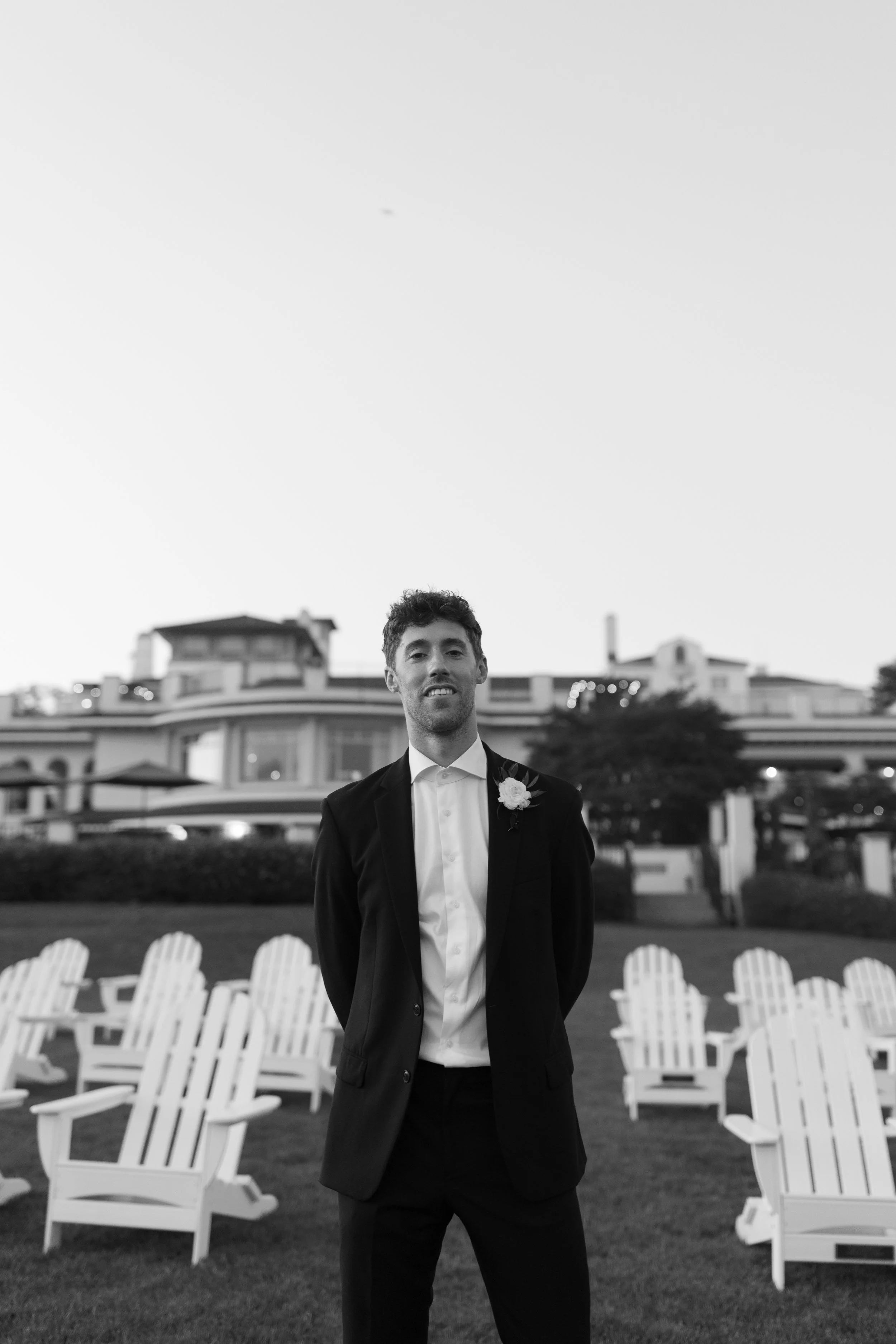 A man in a tuxedo with a white flower boutonniere standing outside in front of a large house with Adirondack chairs arranged in the yard, in black and white.