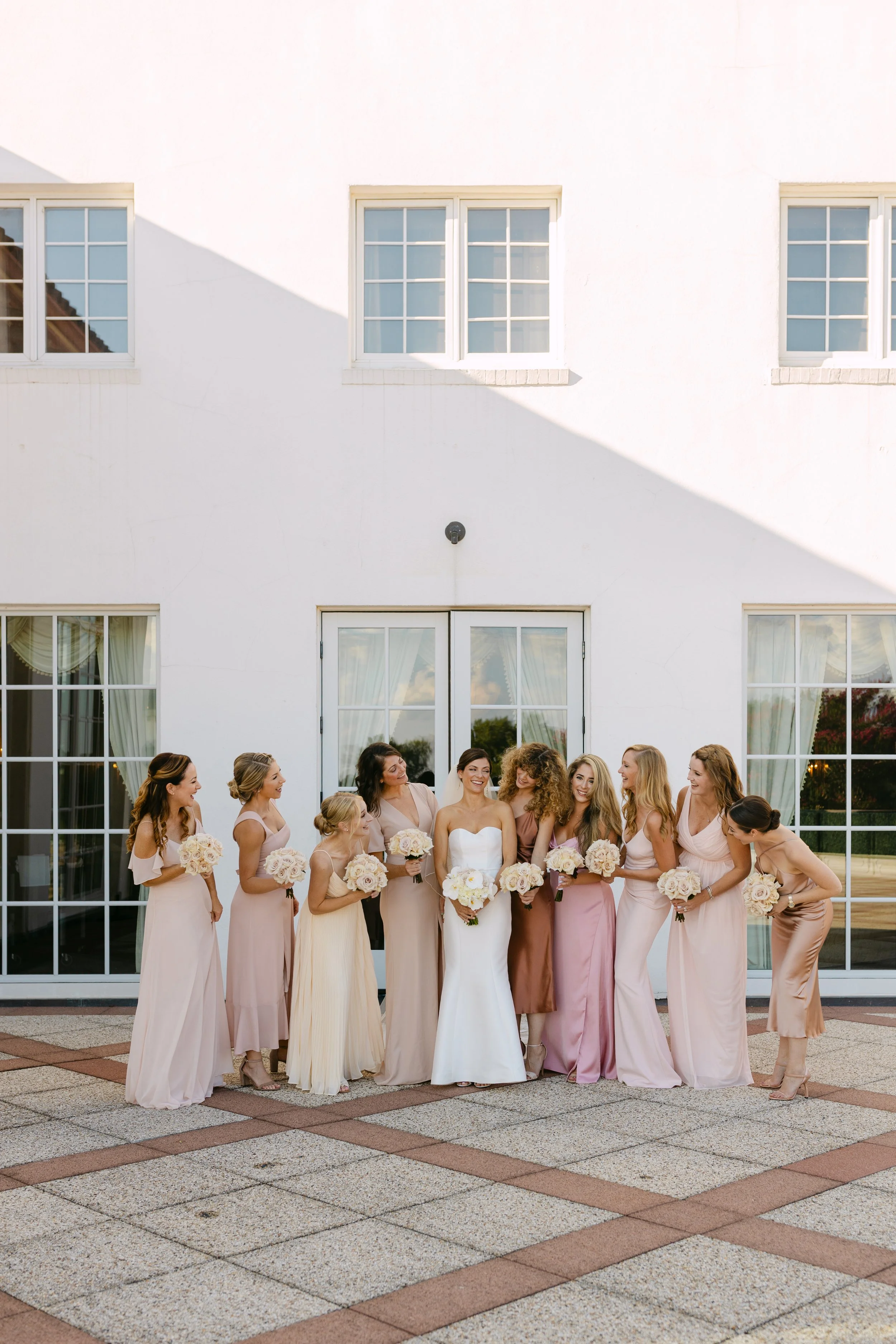 Bride and bridesmaids standing outdoors in front of a white building with large windows, dressed in pastel-colored dresses, holding bouquets, celebrating a wedding.