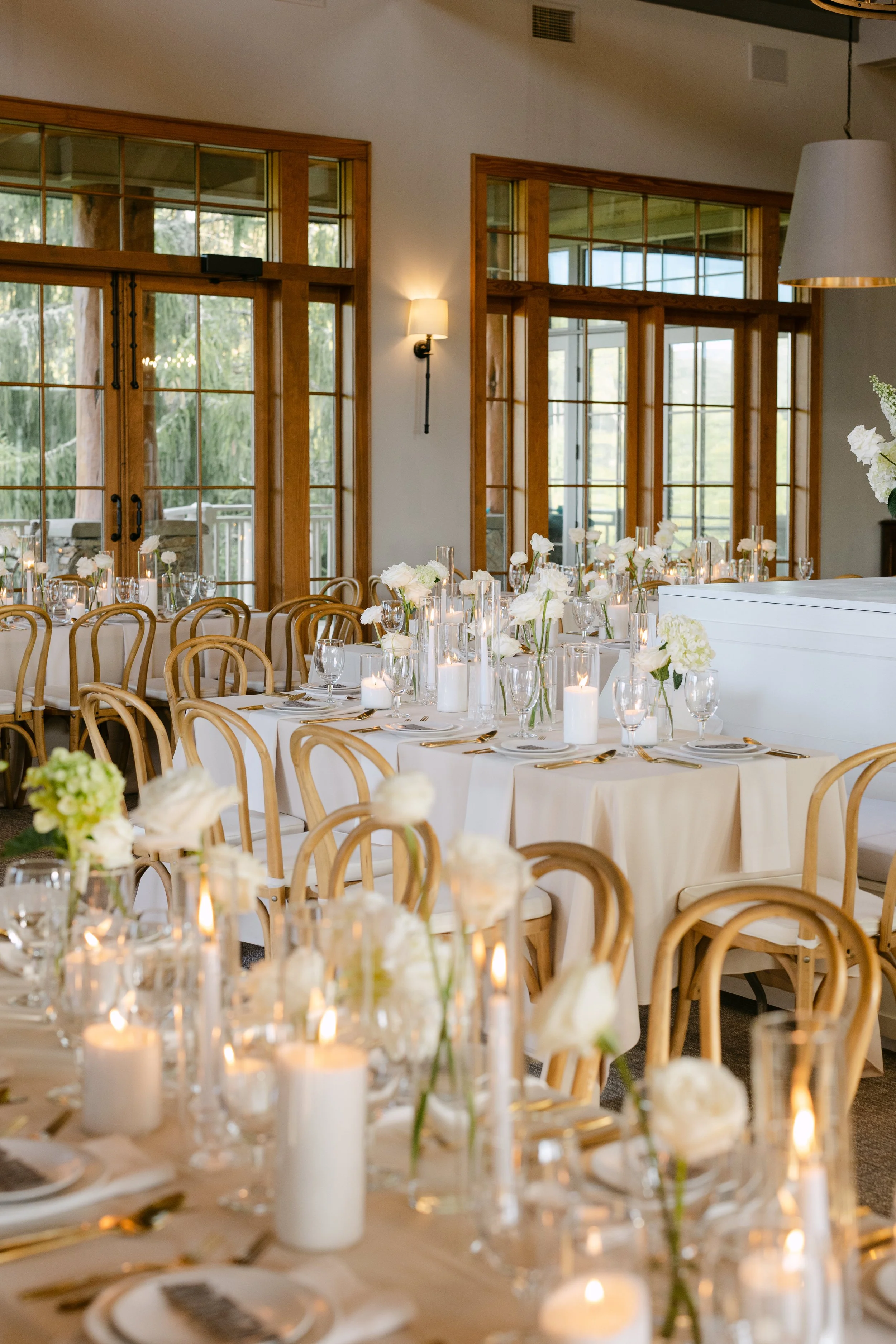 Elegant wedding reception setup with long tables, white tablecloths, floral centerpieces, and candles in a room with large windows and wooden accents.