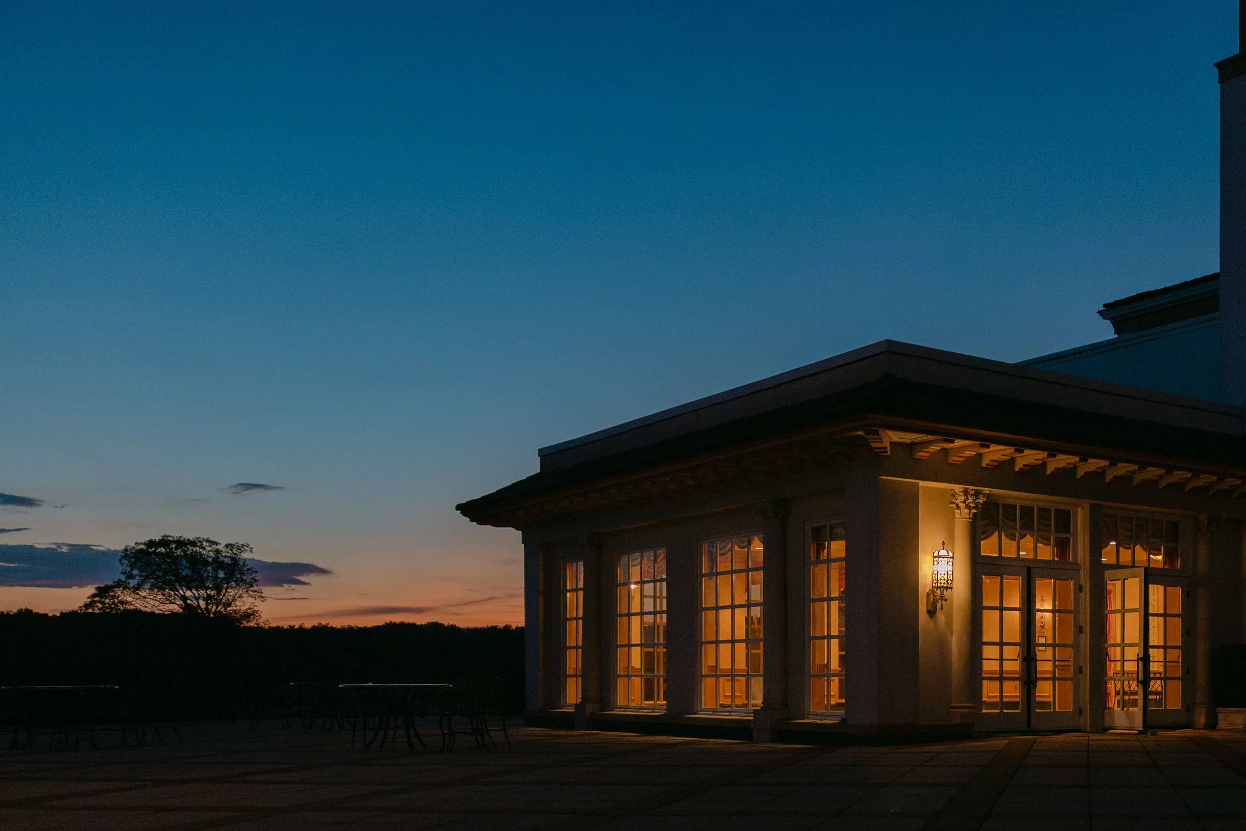 A house at dusk with the interior lights on, large windows, and a porch with outdoor furniture against a darkening sky and silhouetted trees.