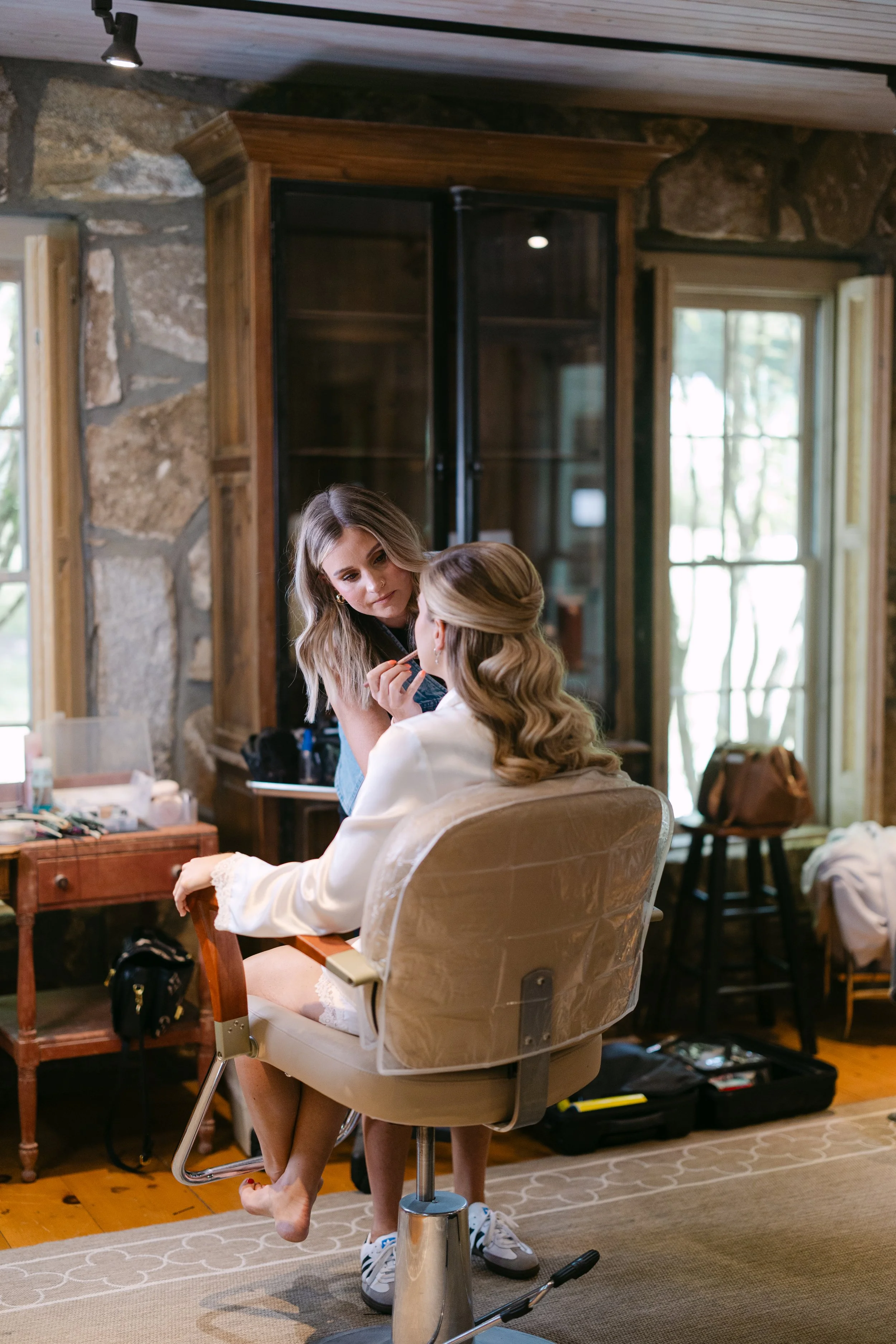 A woman sitting in a chair getting her makeup done by another woman inside a rustic room with stone walls and wooden furniture.