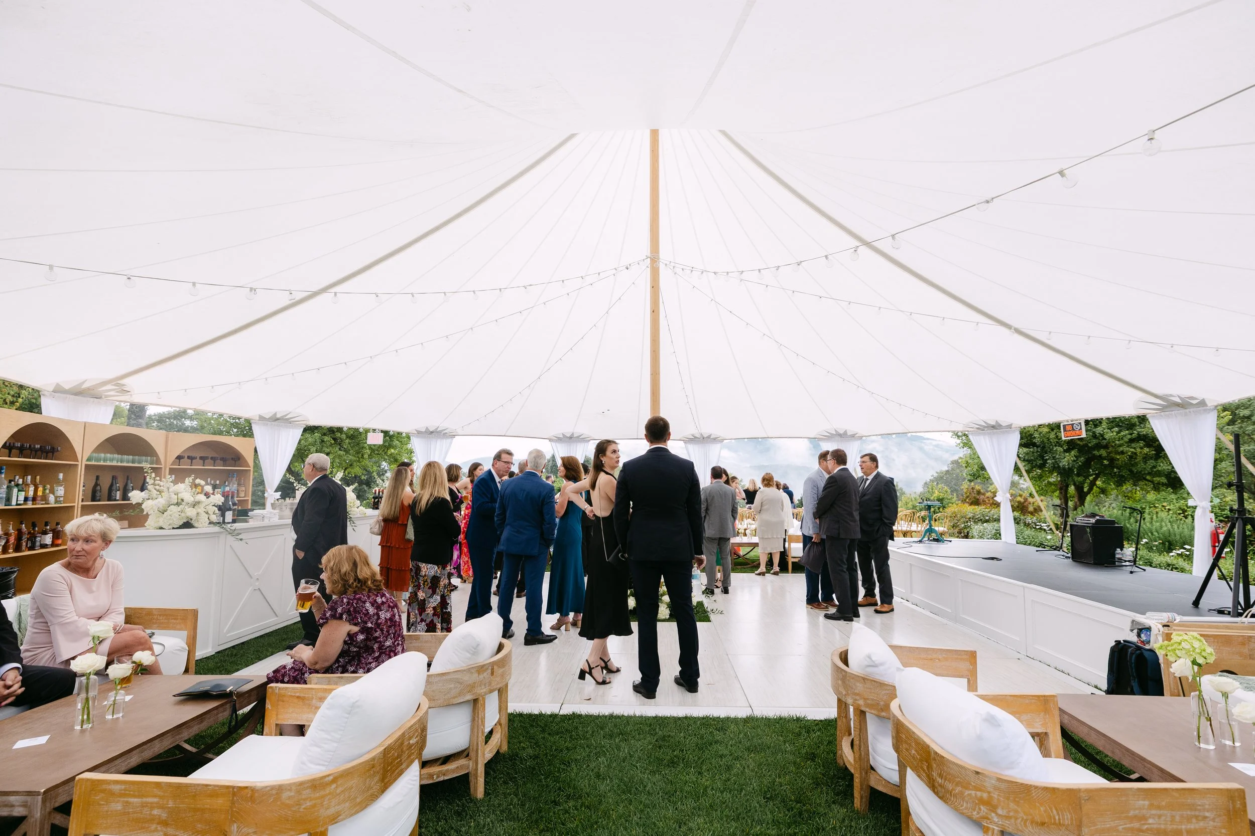 Guests socializing and dancing at an outdoor wedding reception under a large white tent with string lights, with tables and a bar in the background.