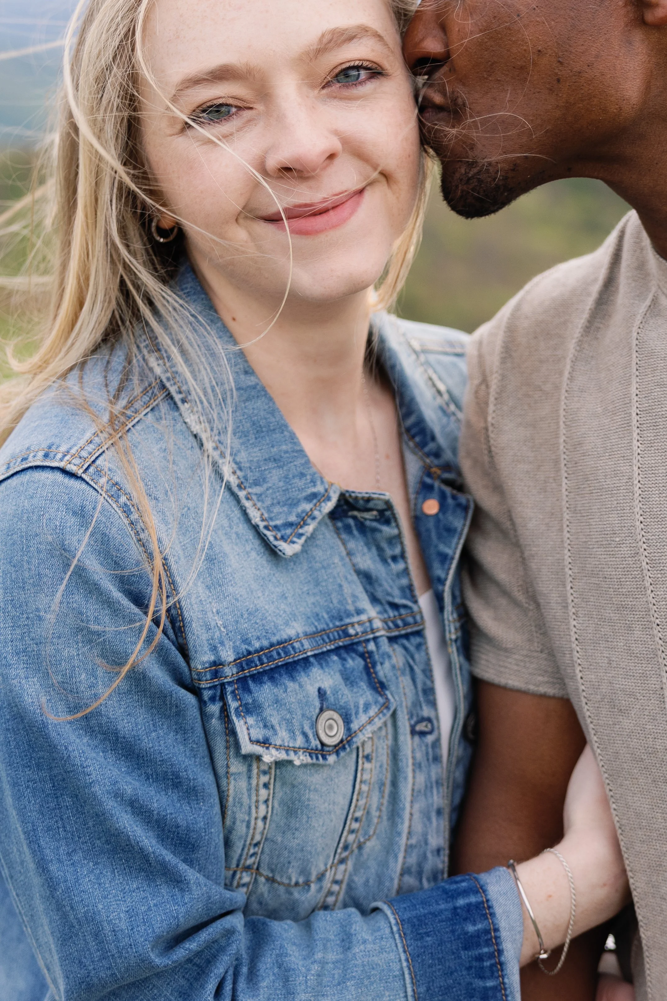 Close-up of two people embracing outdoors, one wearing a denim jacket and the other in a beige shirt, with one person whispering into the other's ear.