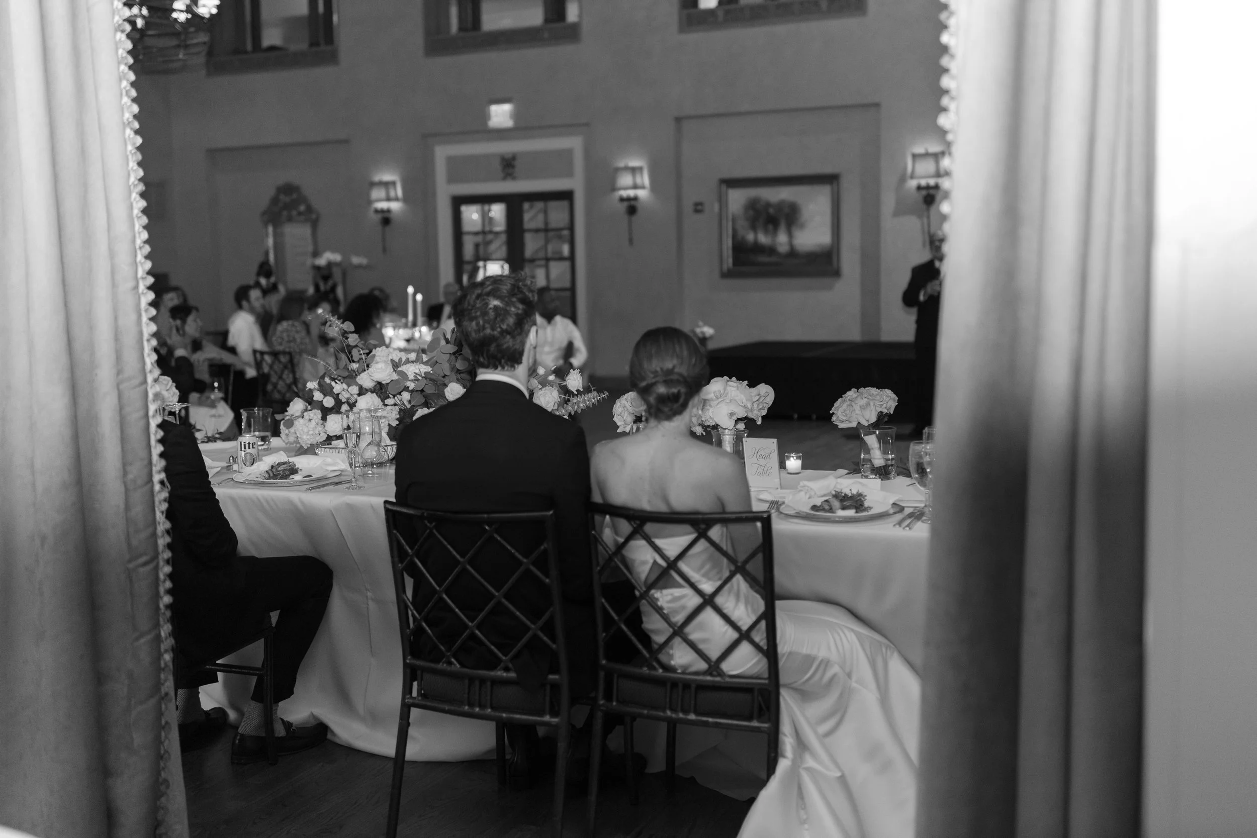 A black and white photo of a wedding reception, seen from behind a curtain, showing a bride and groom sitting at a decorated table, facing a speaker on stage in a banquet hall with guests seated at tables and a painting on the wall.
