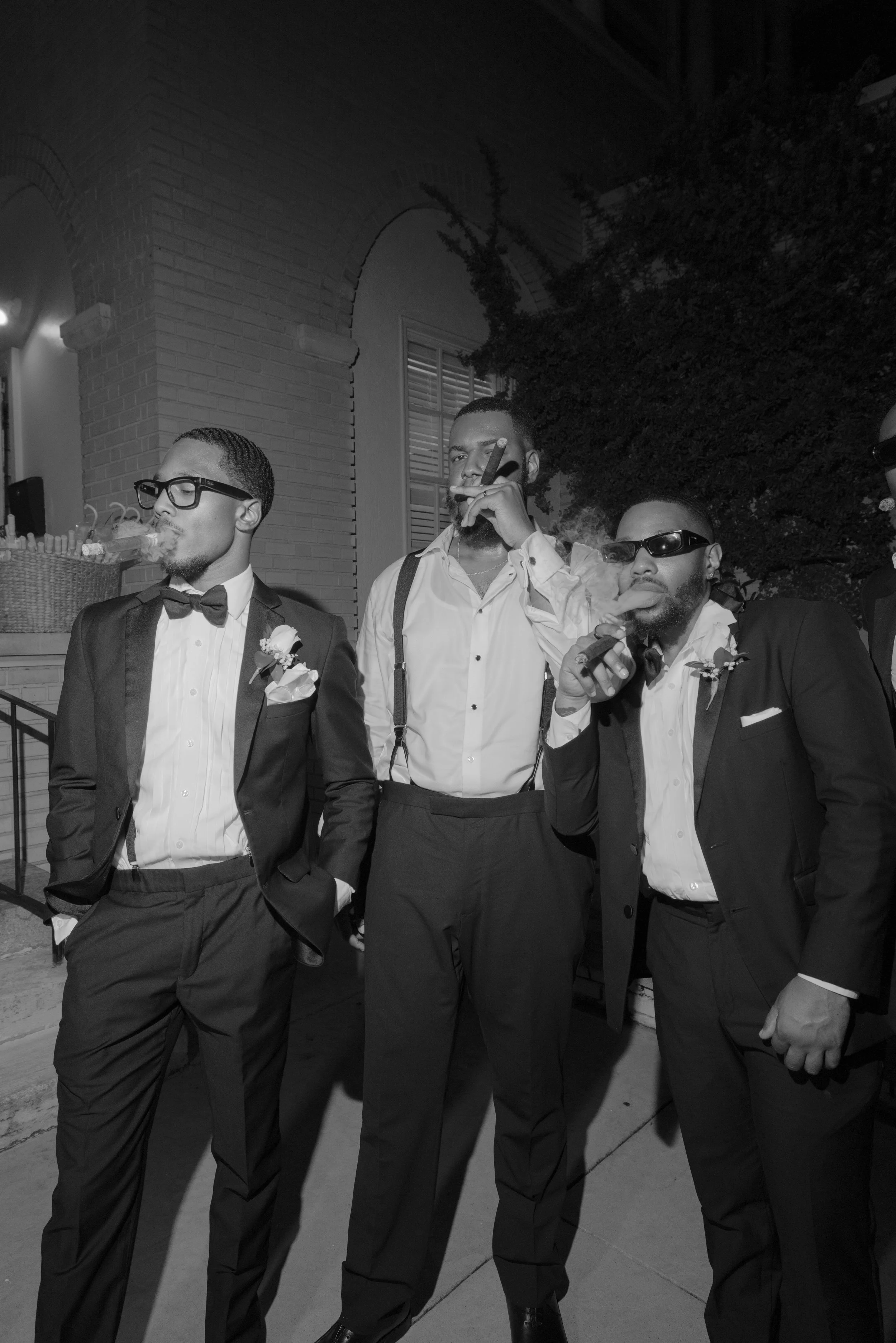 Three men in tuxedos and sunglasses standing outside at night, smoking cigars, with a brick building and bush in the background.