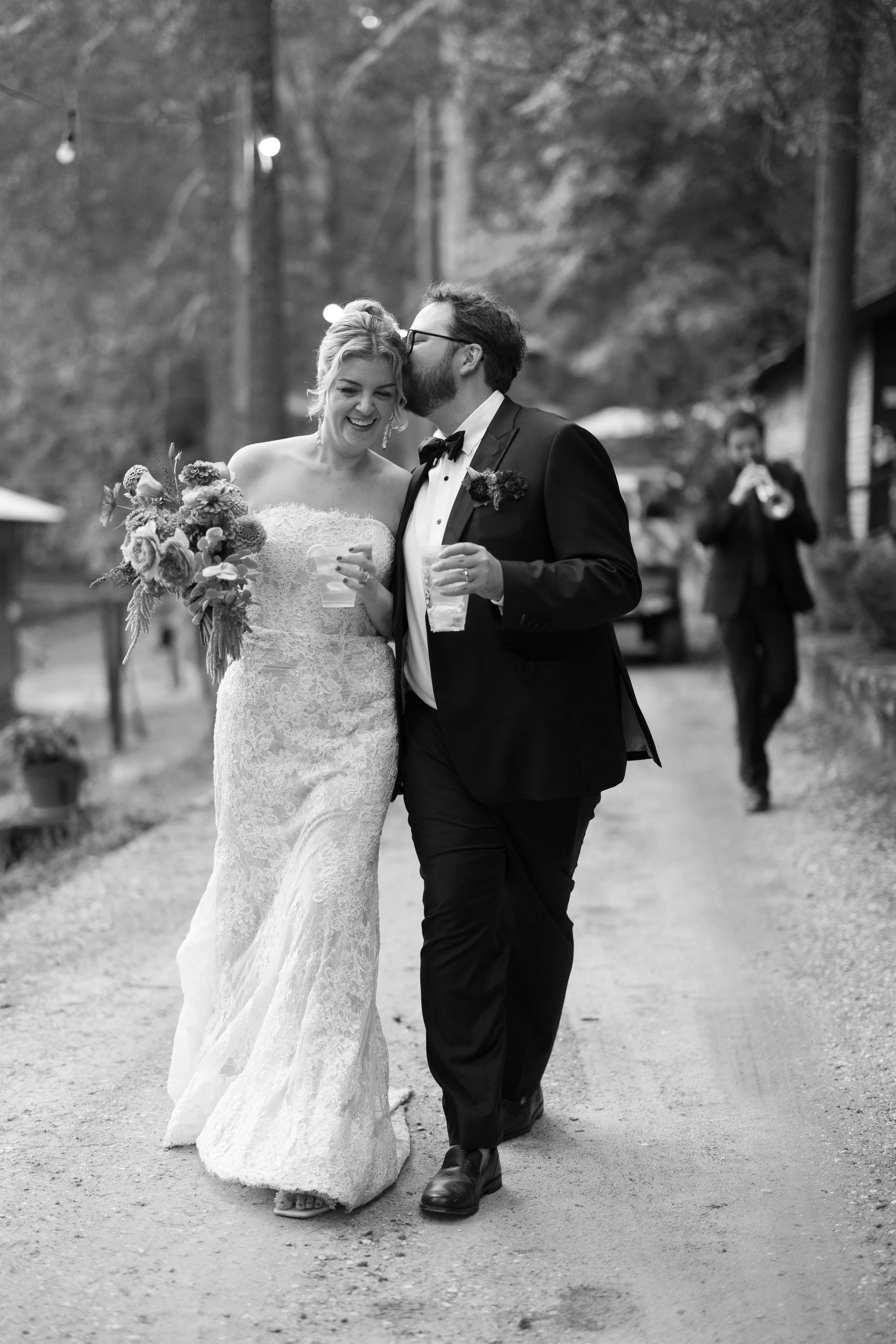 Bride and groom walking outdoors on a gravel path, smiling and sharing a moment, with a man in a tuxedo behind them playing a trumpet.