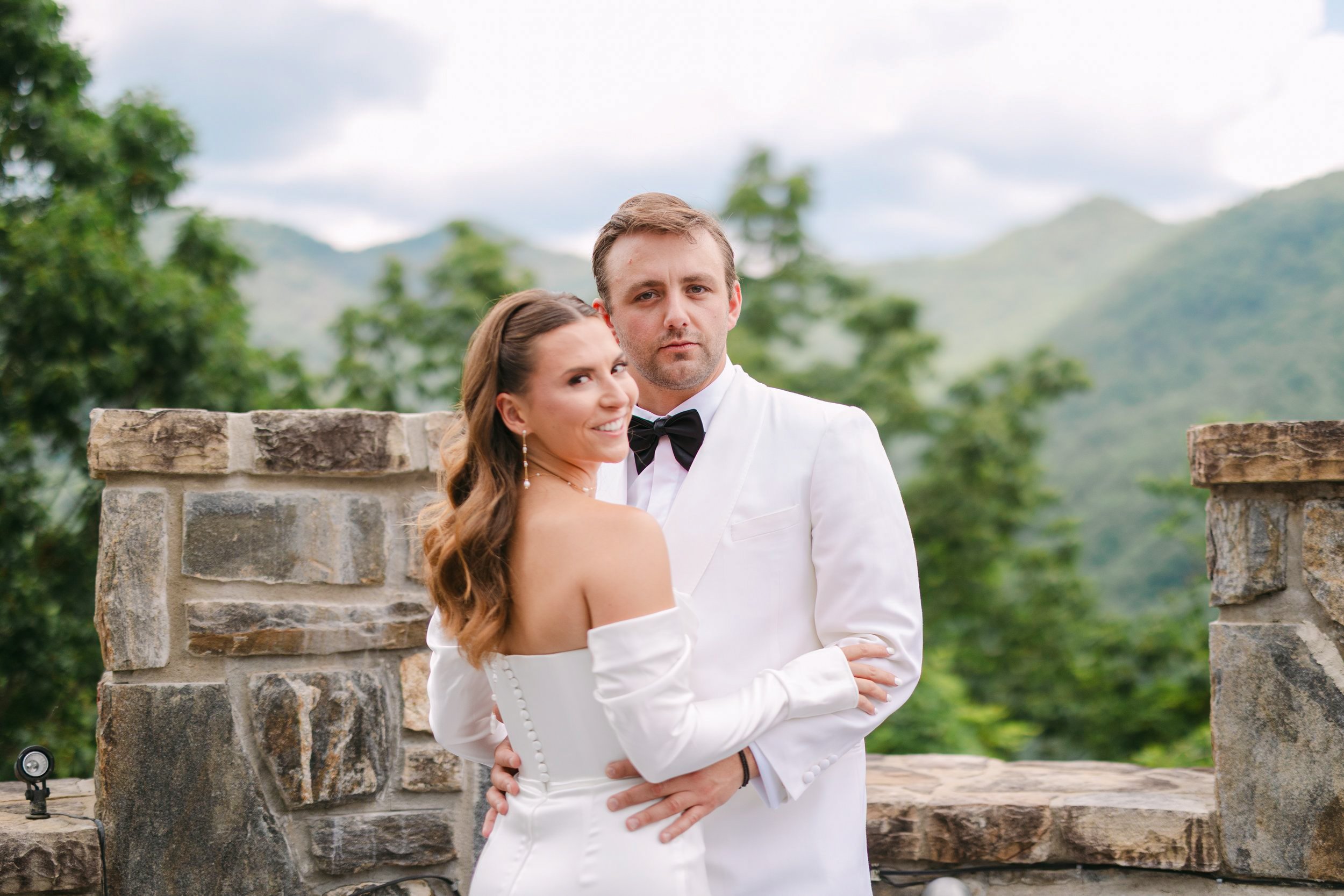 A newlywed couple dressed in wedding attire stands outdoors with a stone wall and a scenic mountain landscape in the background. The bride is smiling and looking at the camera, while the groom is looking slightly serious, with his arm around the brid
