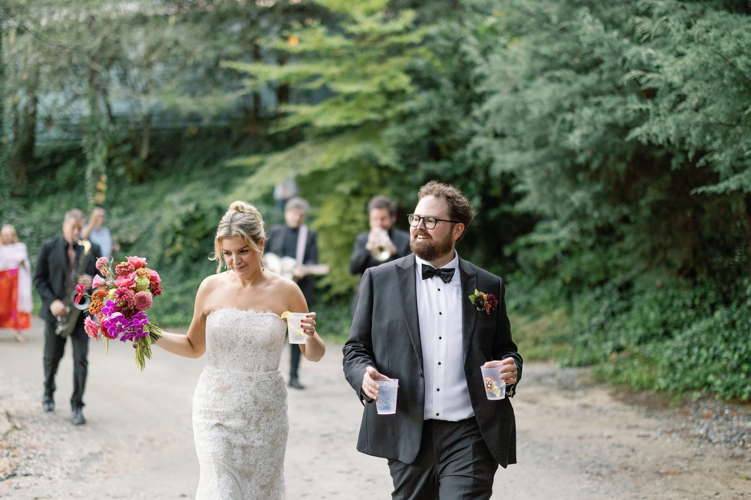 Bride in a white lace strapless wedding dress holding a bouquet of pink and purple flowers, and groom in a black tuxedo with a bow tie, walking outdoors with drinks in hand, with guests and greenery in the background.