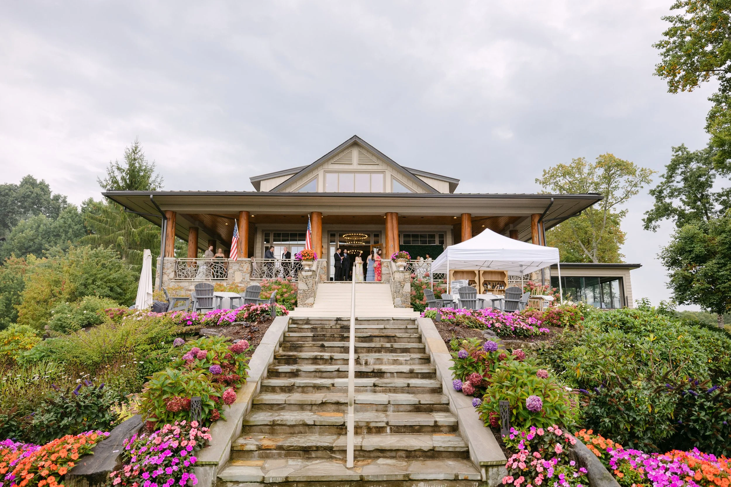 A wedding reception on a large outdoor terrace of a house, with guests mingling, flower arrangements, and a white canopy tent.