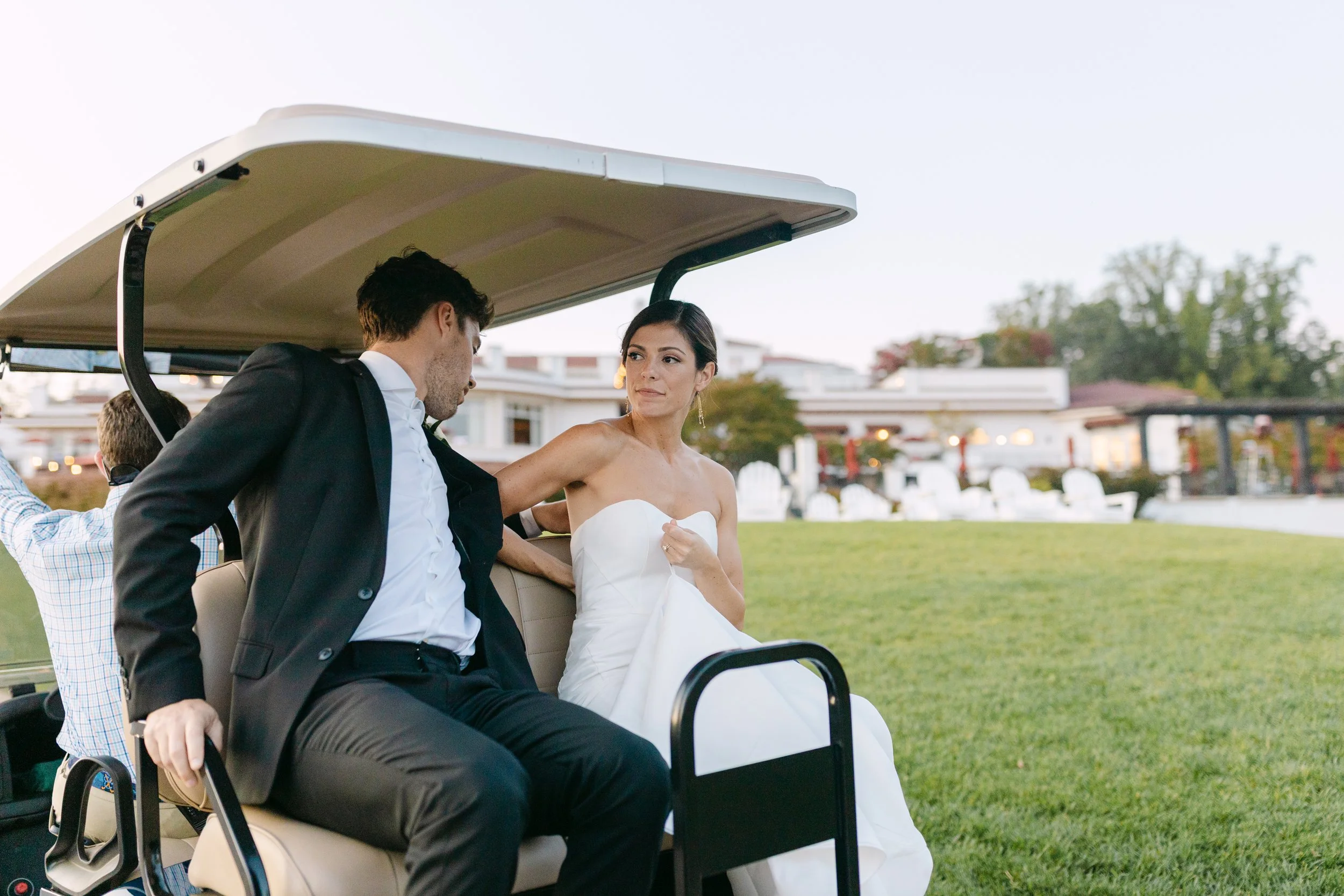 A bride and groom sitting on a golf cart outside, with the bride in a strapless white wedding gown and the groom in a black suit, while a person driving the golf cart is partially visible in the background.
