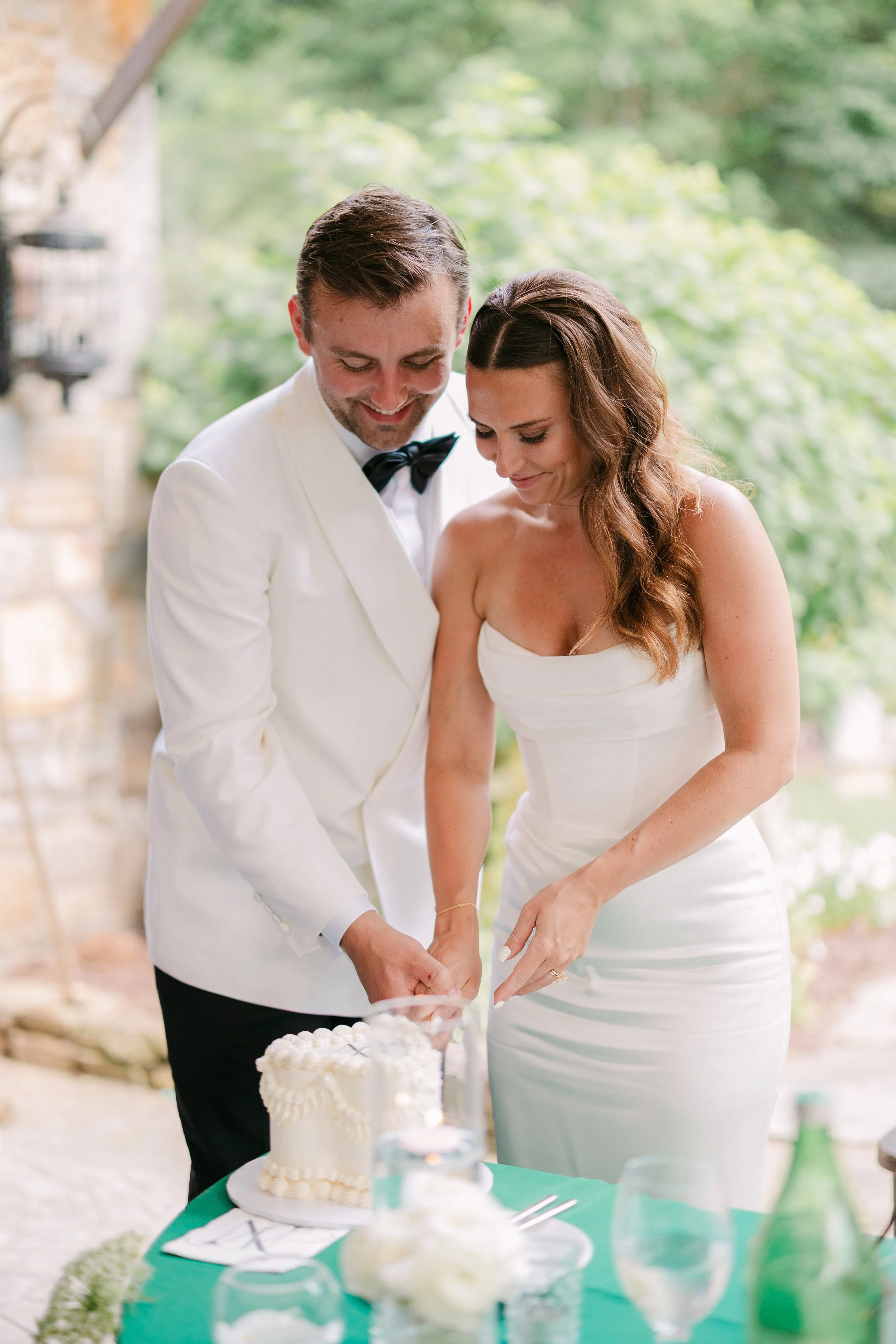 A newlywed couple, dressed in wedding attire, is cutting their wedding cake together outdoors. The groom is wearing a white tuxedo jacket with a black bow tie, and the bride is wearing a strapless white wedding gown with wavy brown hair. They are smi