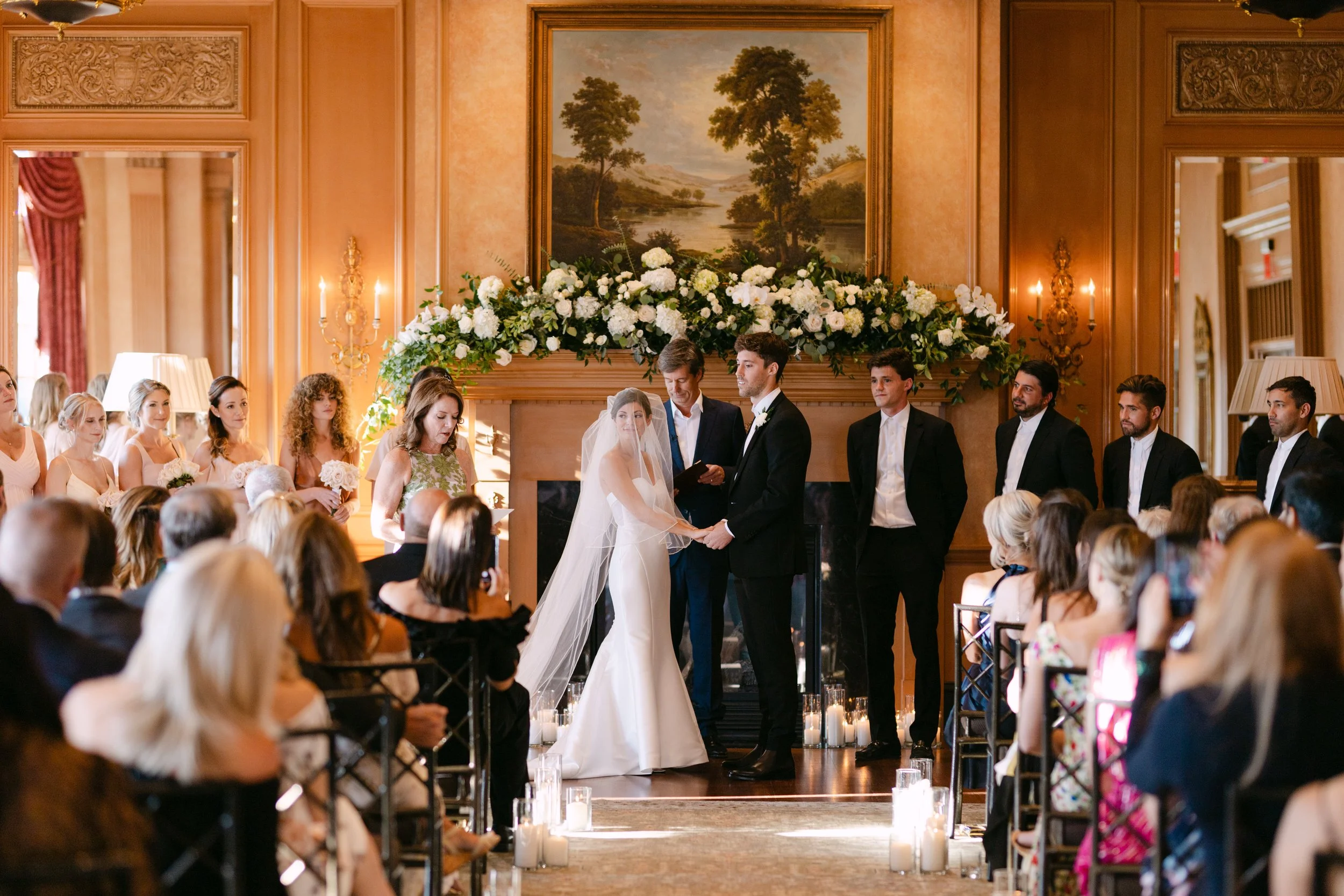 A wedding ceremony taking place indoors with a couple holding hands in front of officiant, surrounded by guests. The bride is in a white gown with a veil, and the groom in a dark suit and bowtie. The room is decorated with flowers and a large landsca