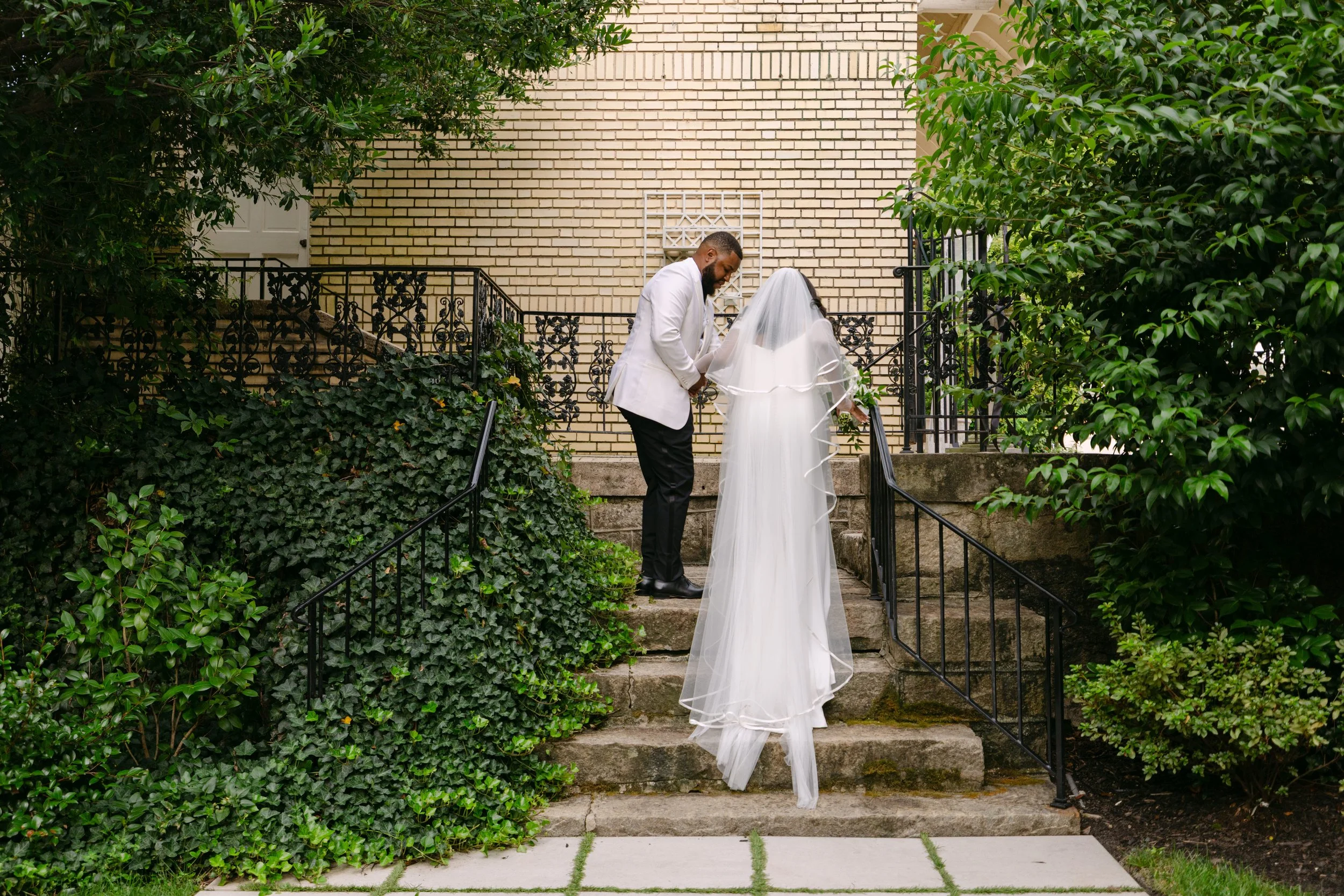 A bride and groom holding hands while standing on outdoor stone steps, with lush green bushes and trees on either side, and a brick building with a white door in the background.