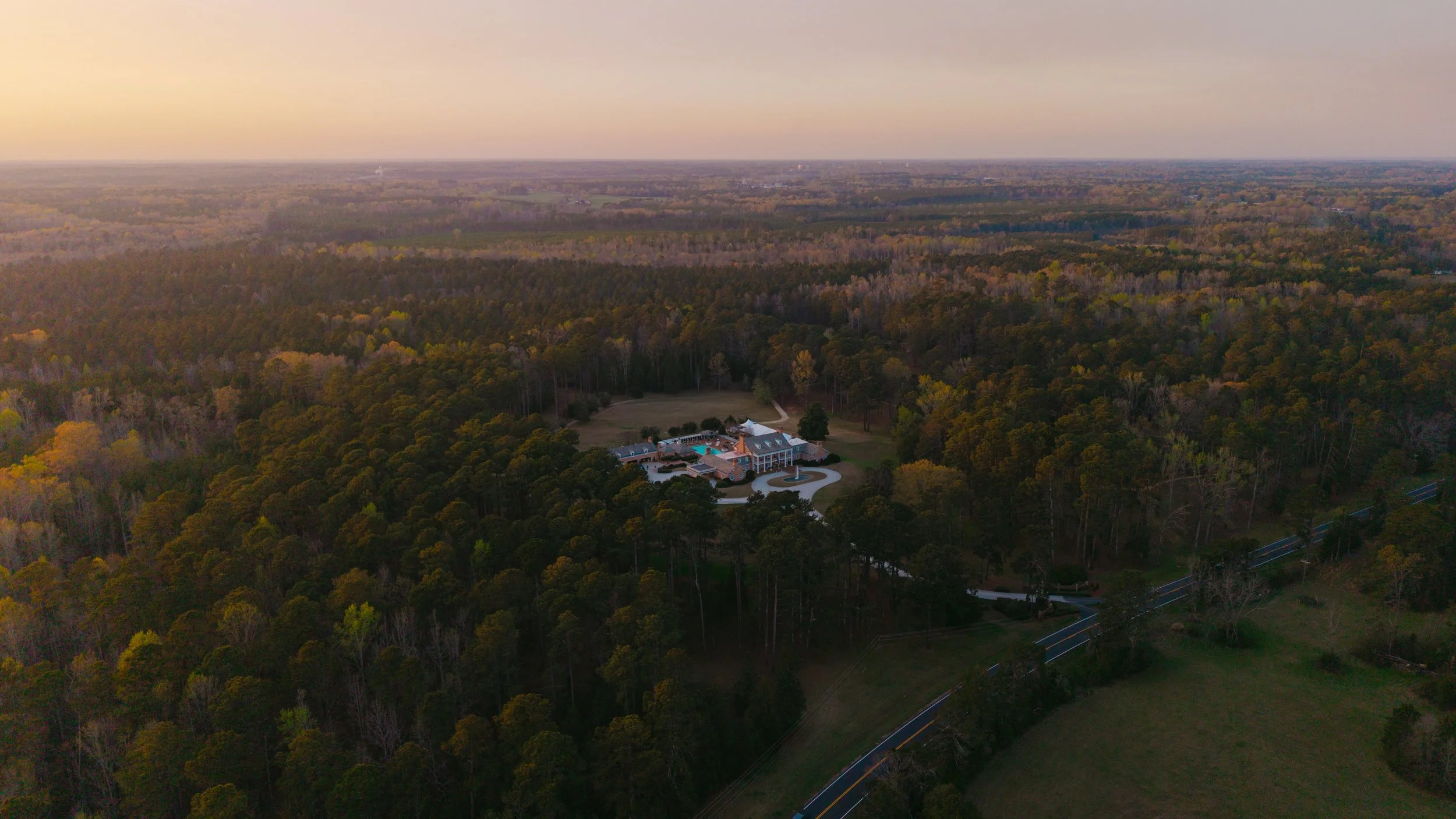 Aerial view of a large mansion surrounded by trees, with a driveway leading to the entrance, during sunset.