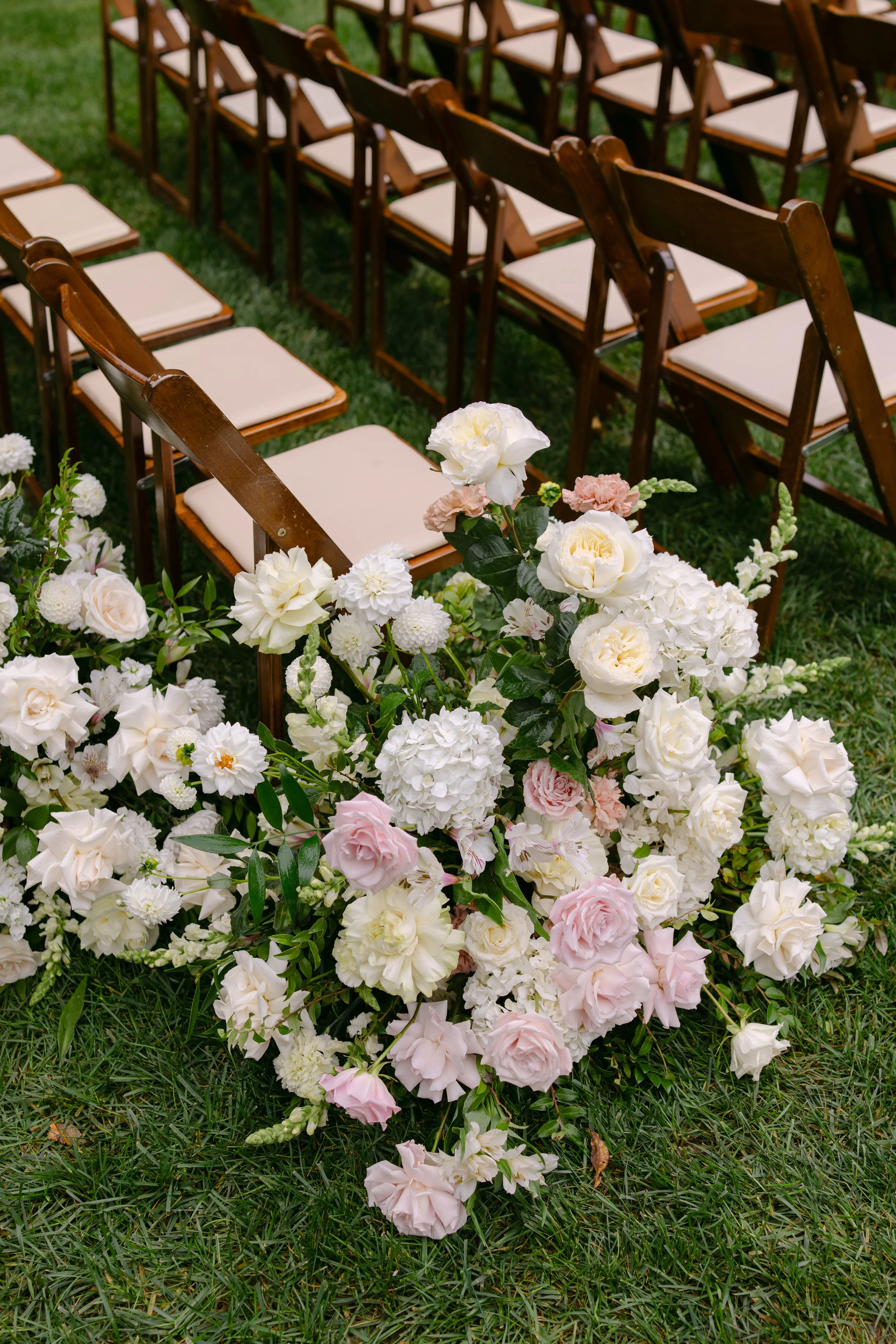Wooden folding chairs arranged on grass behind a lush floral wedding arrangement of white and blush pink flowers